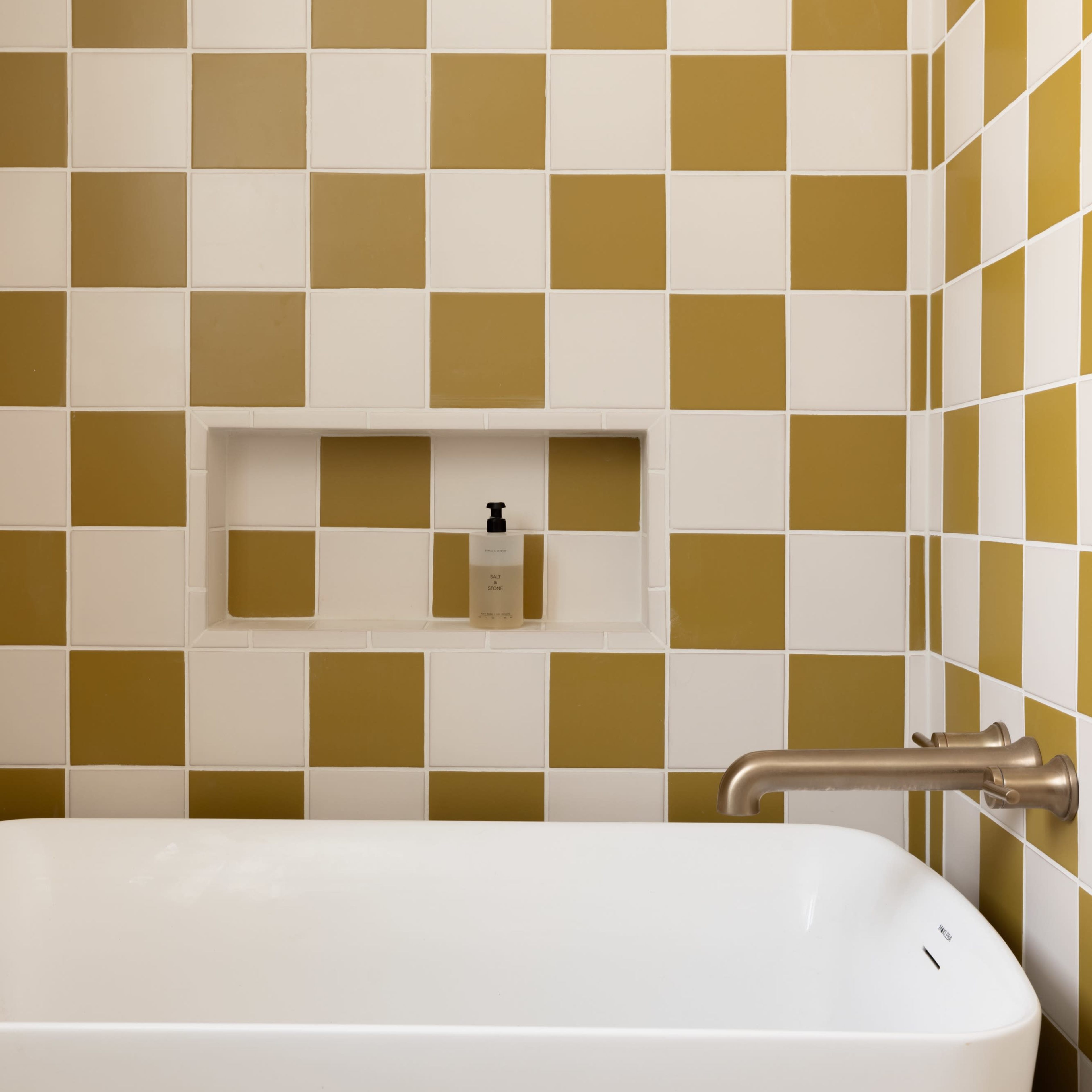 A modern bathroom with a white bathtub and a wall featuring alternating white and mustard-colored checkered tiles.