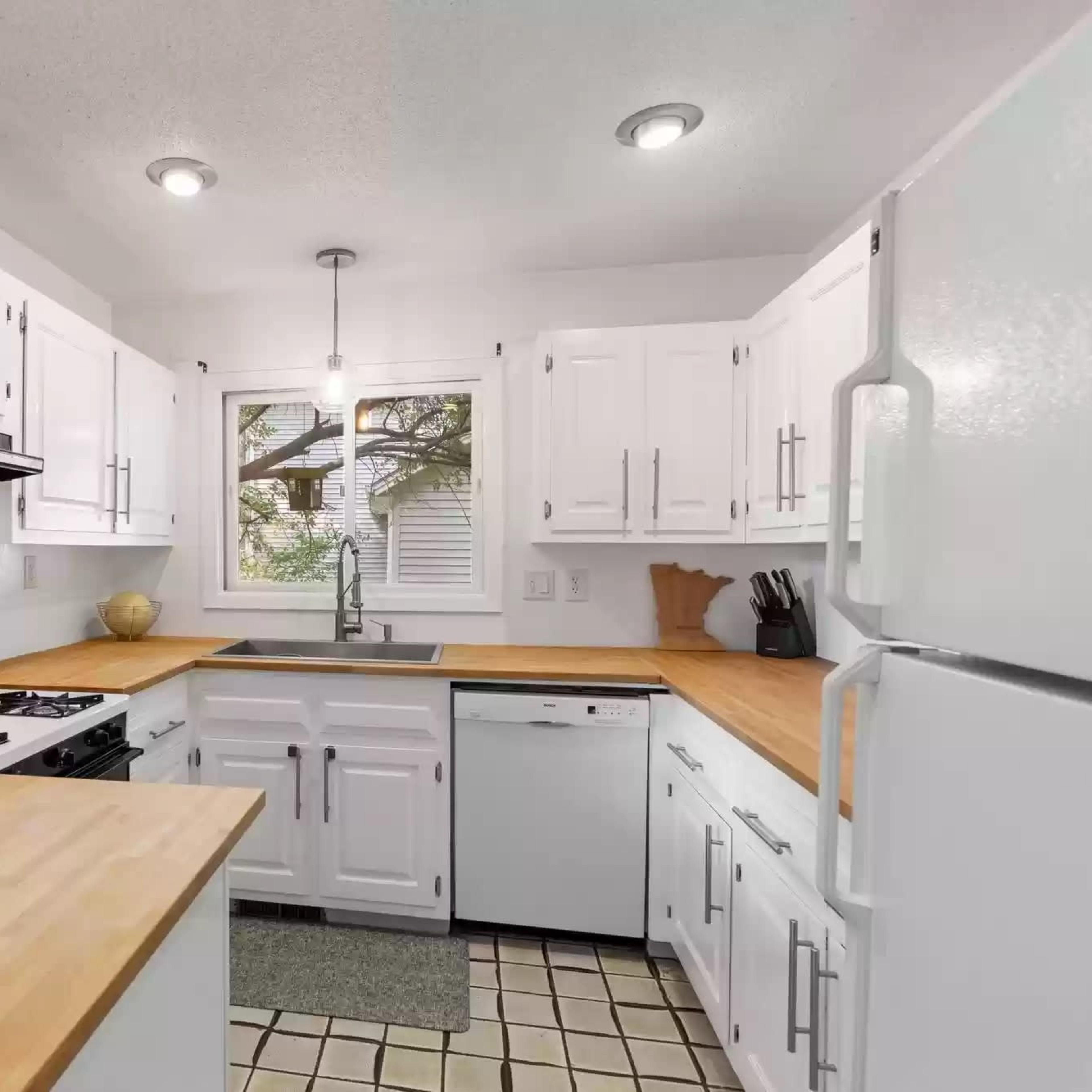 A compact kitchen with white cabinets and countertops, equipped with a gas stove, dishwasher, and a window offering a view of greenery outside.