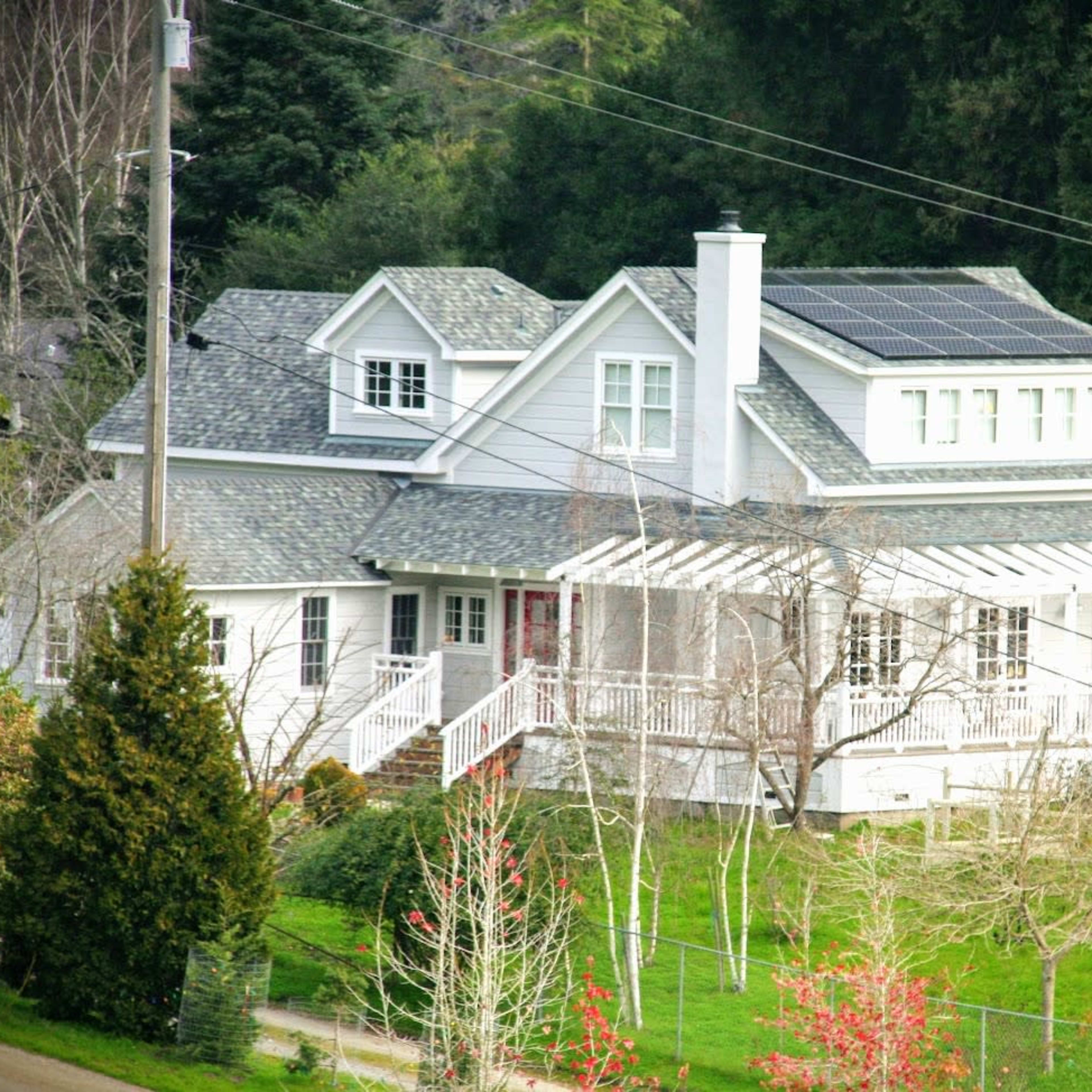 A two-story house with a gabled roof and solar panels is surrounded by trees and has a front porch with a railing.