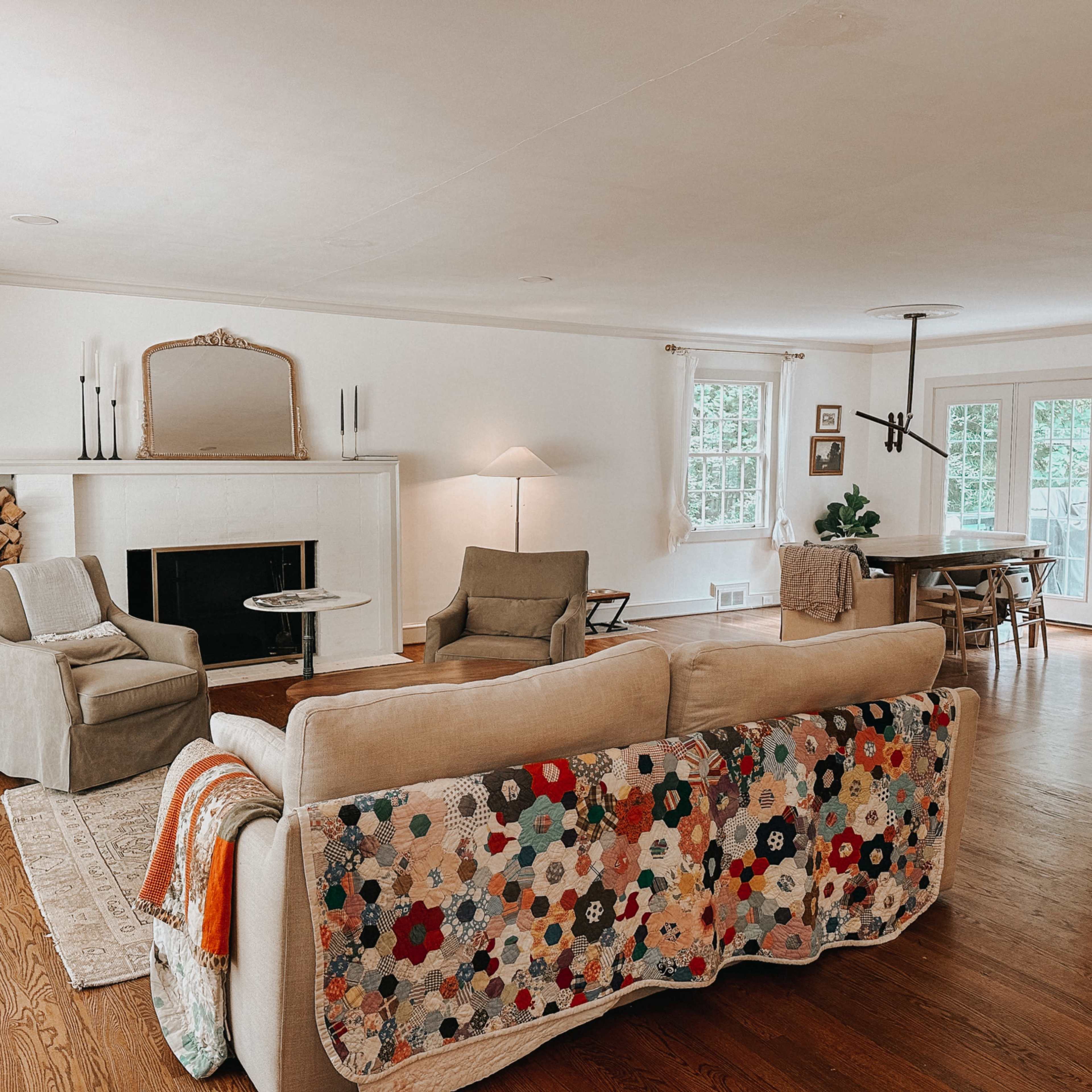 A living room with a sofa covered in a colorful quilt, two chairs, a fireplace with stacked wood, and a dining area in the background.