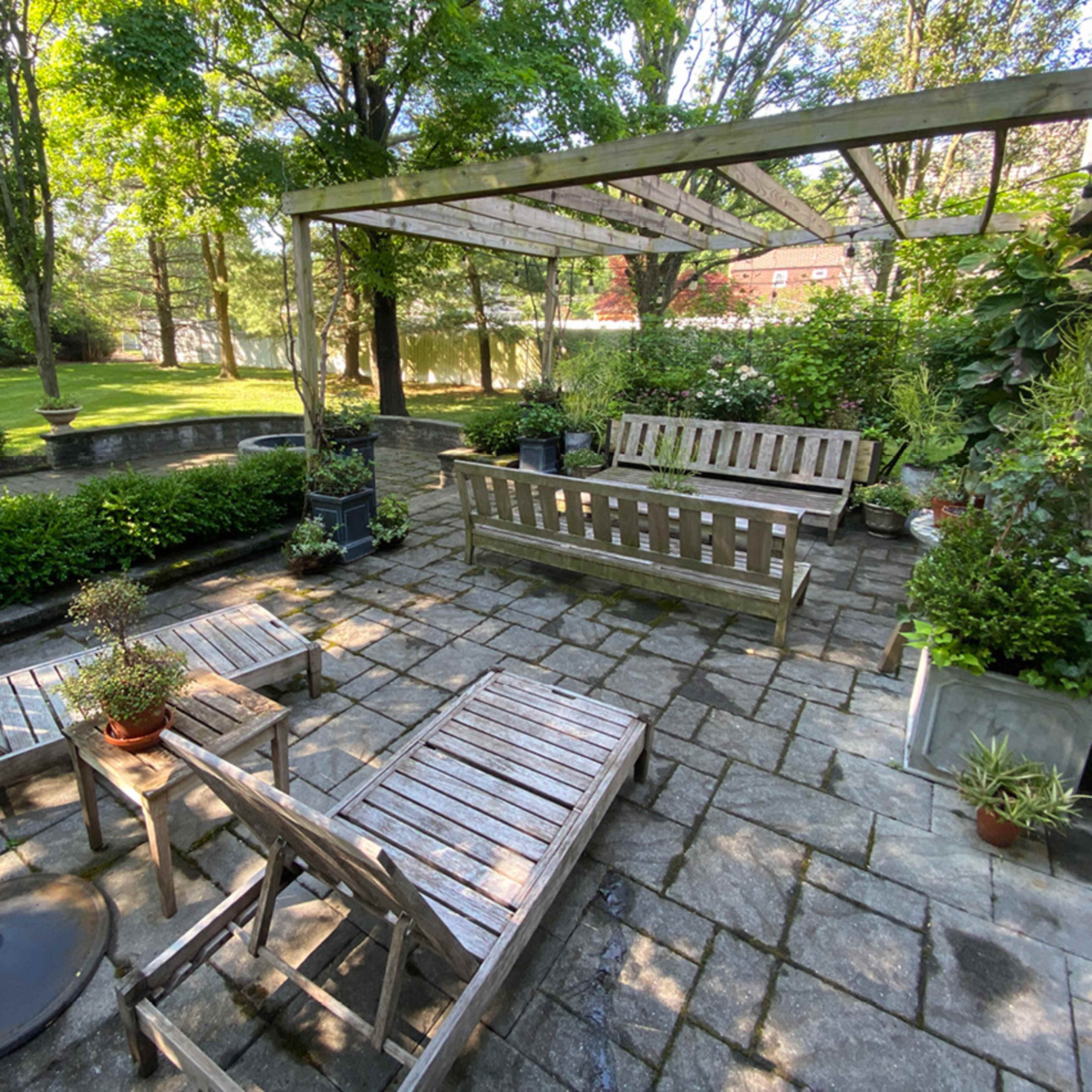 A stone patio features wooden furniture and potted plants under a wooden pergola in a shaded outdoor space.