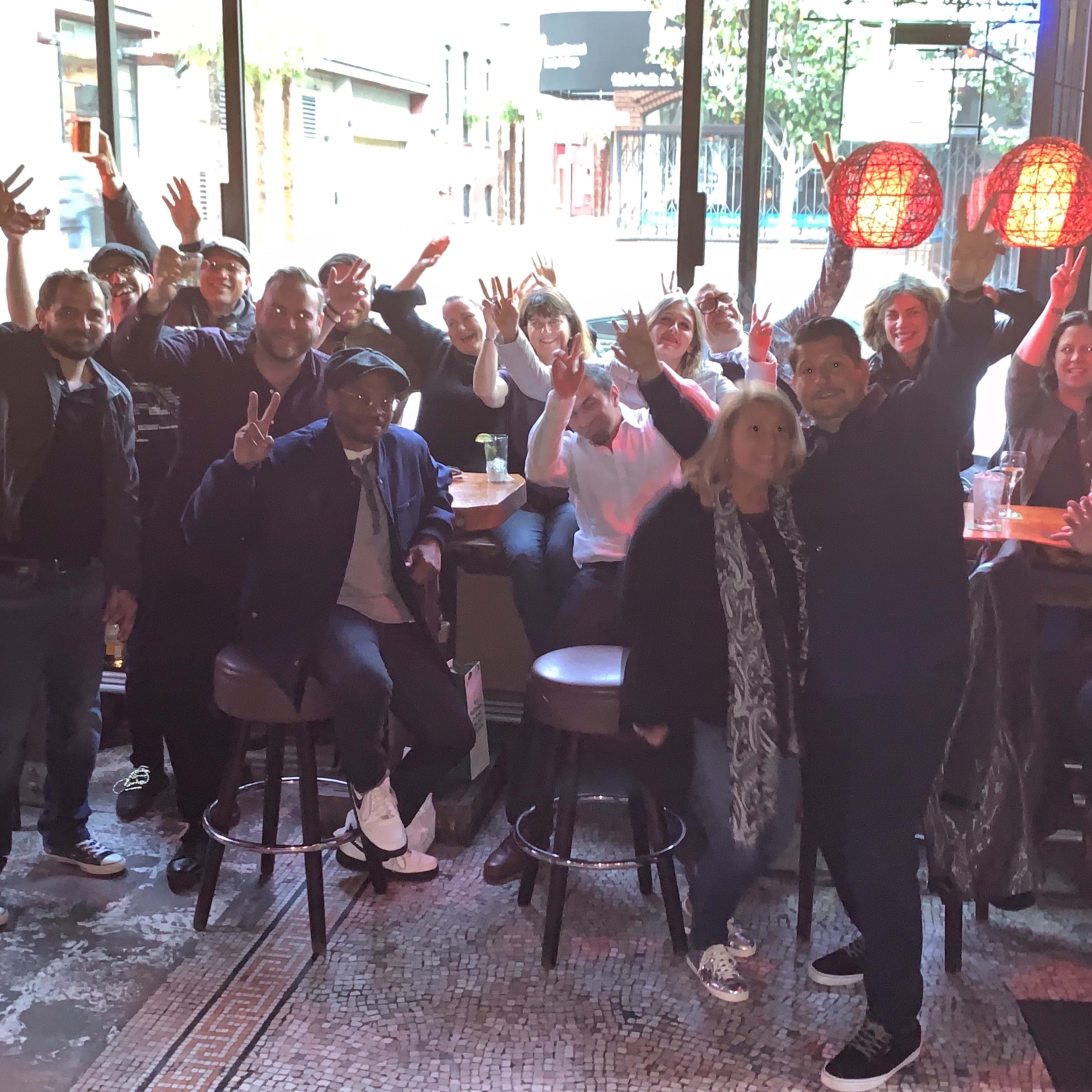 A group of people poses for a photo with their hands raised in a bar setting, surrounded by tall windows and hanging red light fixtures.