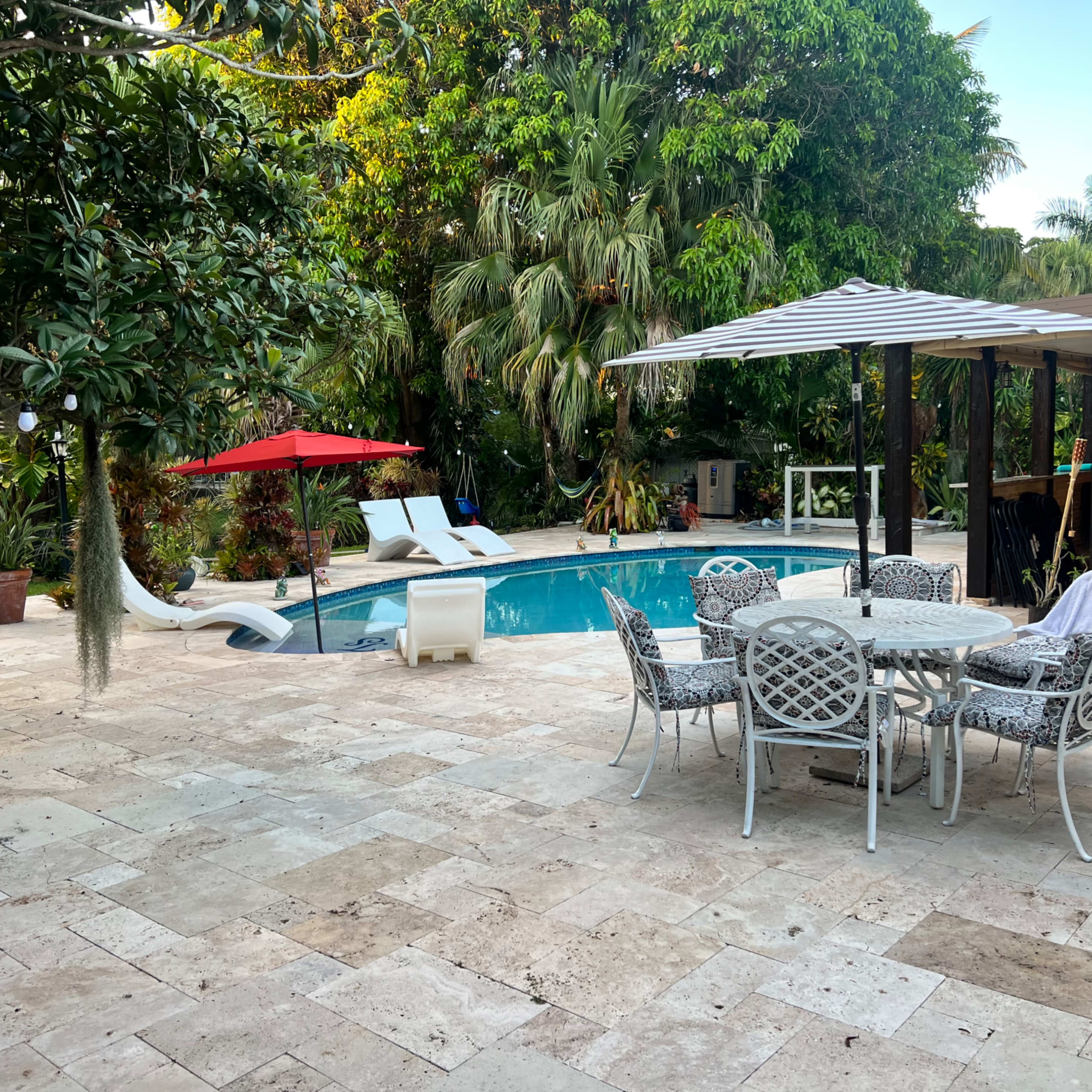 A patio area with a dining table and chairs adjacent to a swimming pool, surrounded by lush tropical vegetation and a shaded seating area.