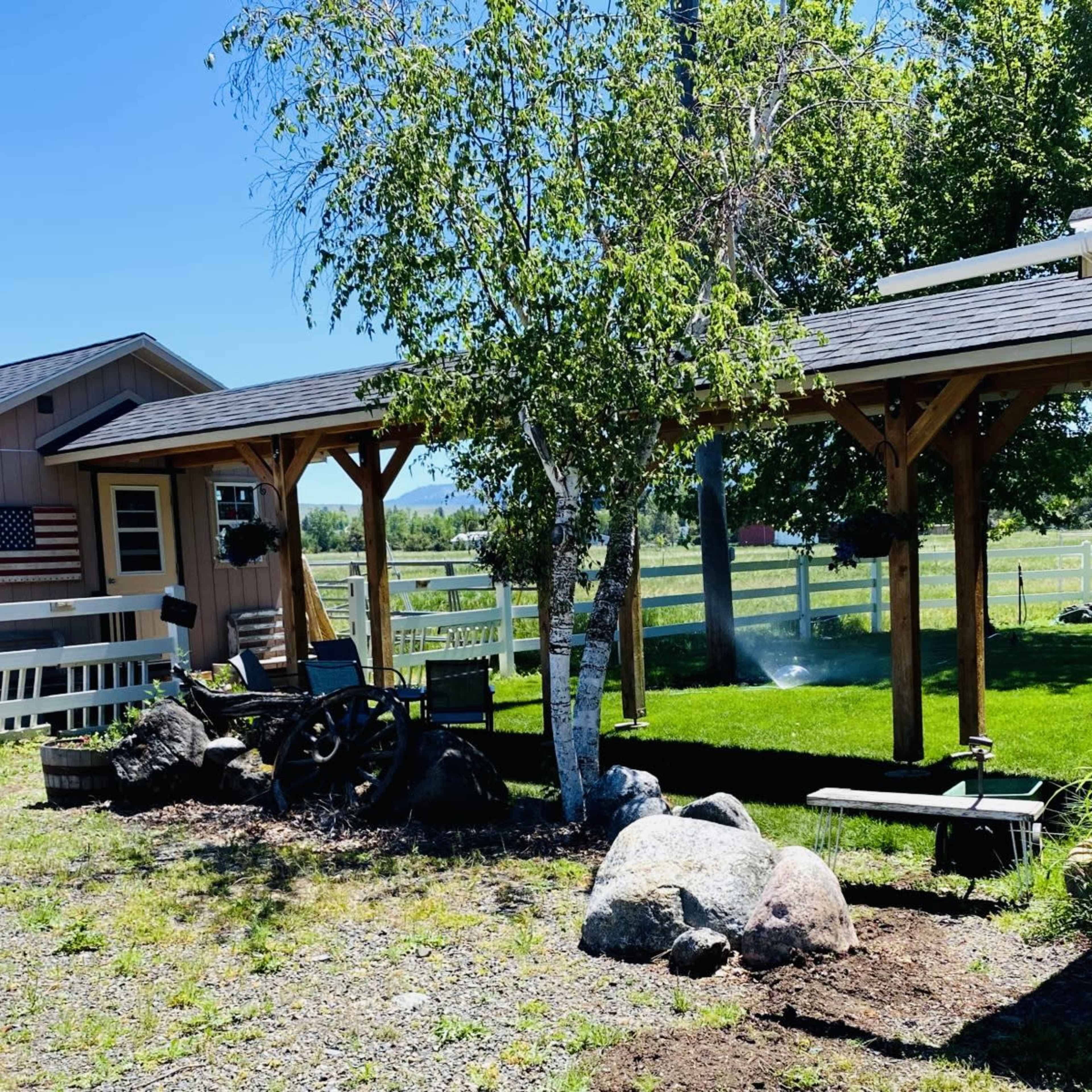 A small cabin with an American flag and a shaded seating area is set among rocks and greenery in a rural landscape.