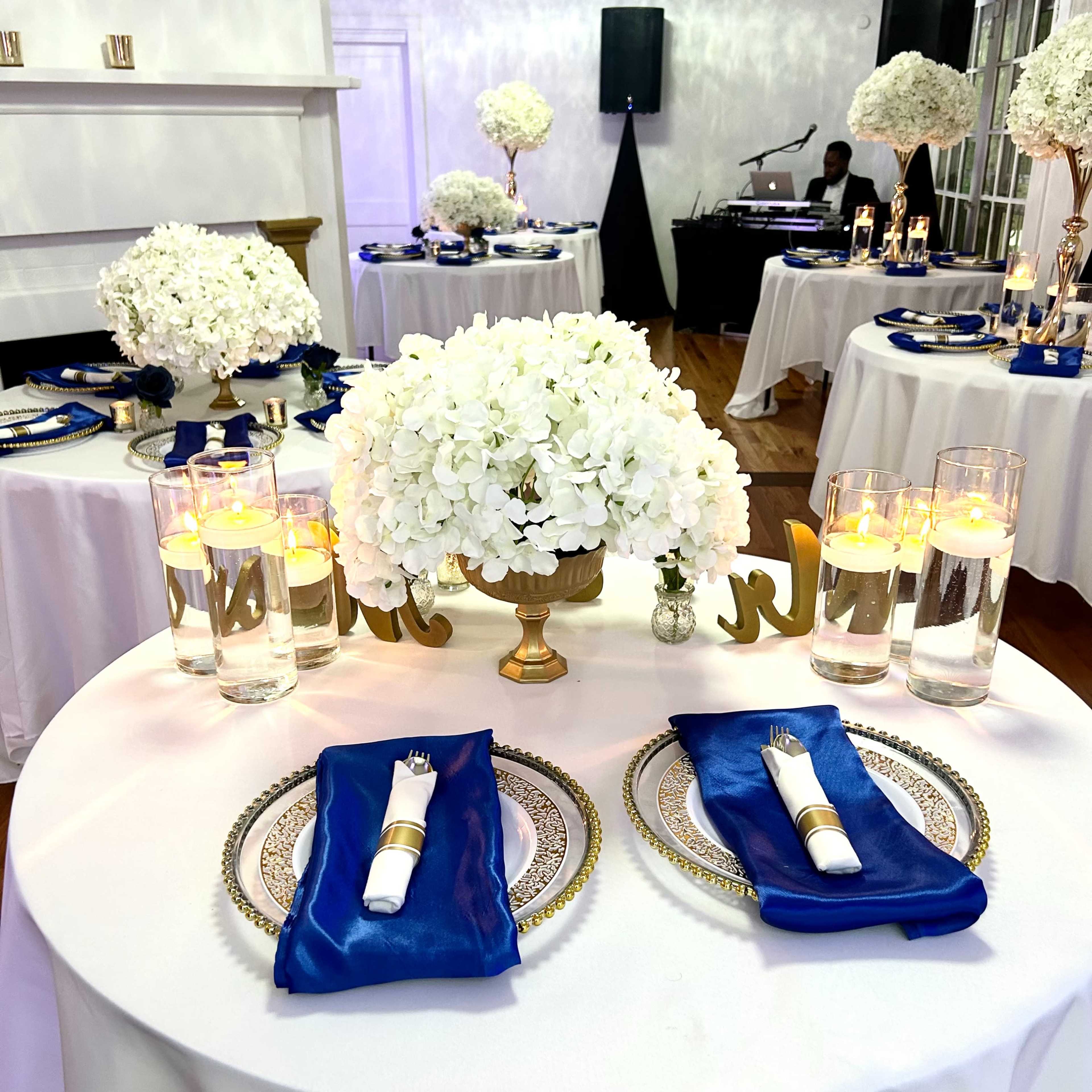 A round table is set with white hydrangeas, blue napkins, elegant glassware, and decorative gold accents in a banquet-style room.