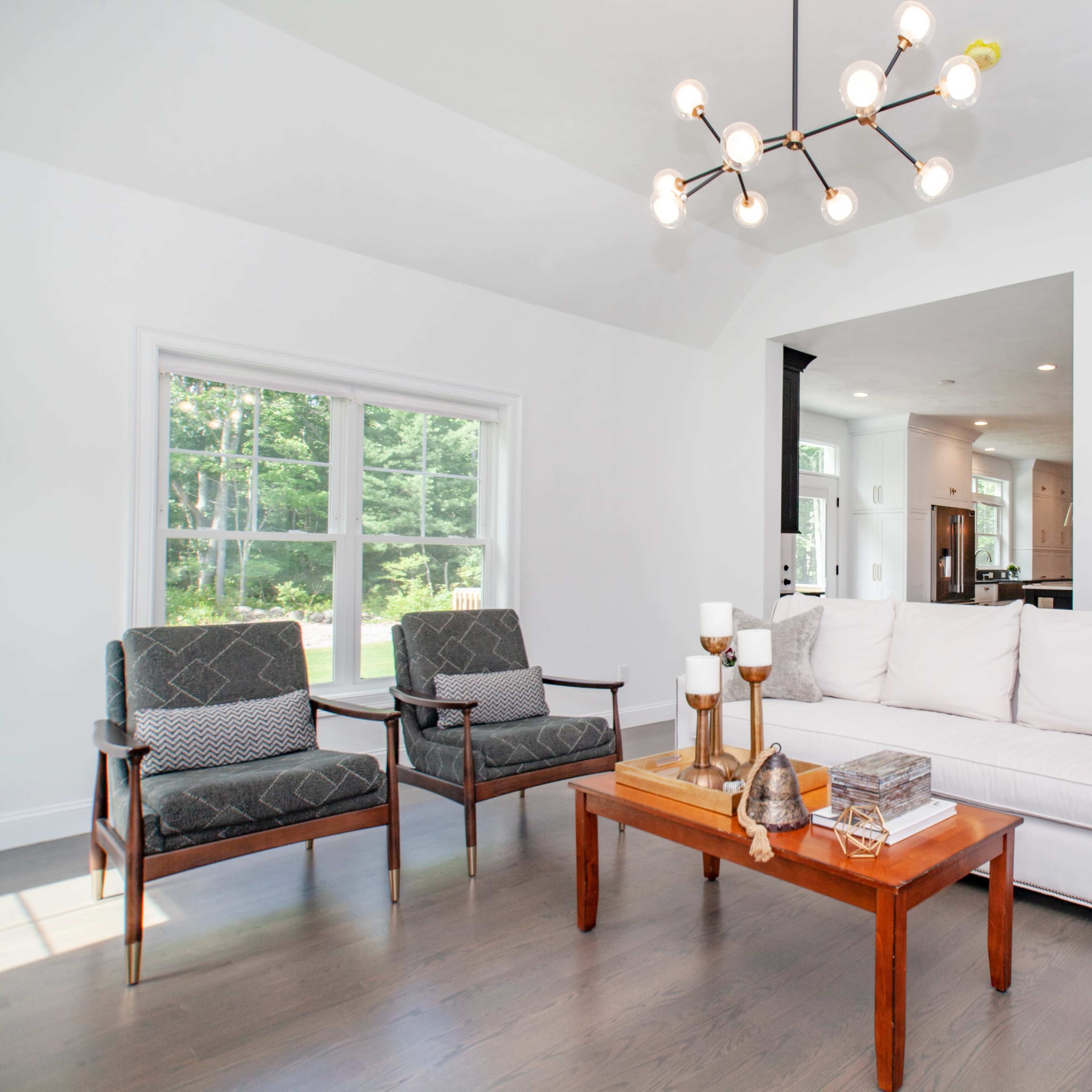 A well-lit living room featuring a white sectional sofa, two armchairs, a wooden coffee table, and a large window with a view of greenery outside.