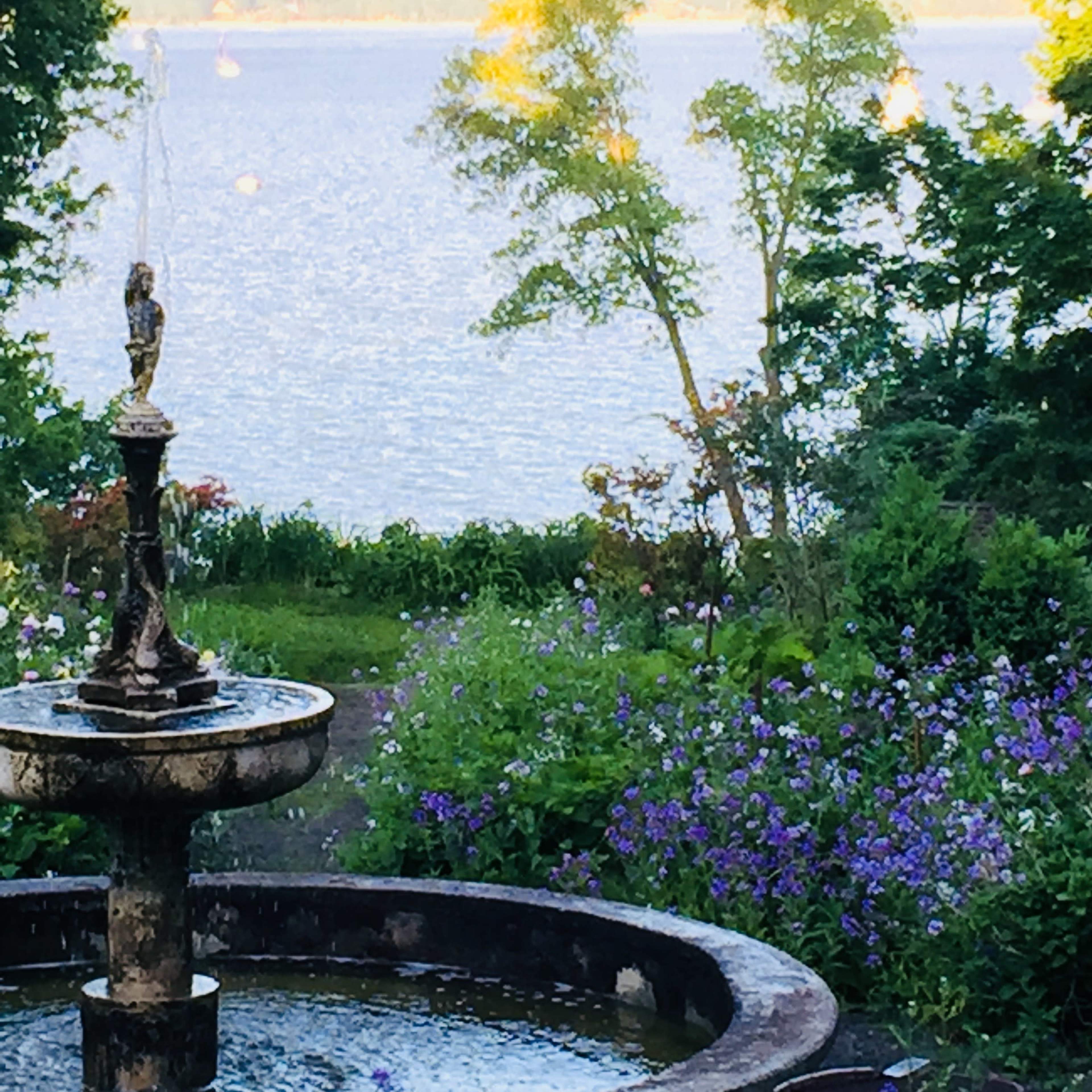 A stone fountain sits in a garden filled with flowers, overlooking a calm lake surrounded by trees.