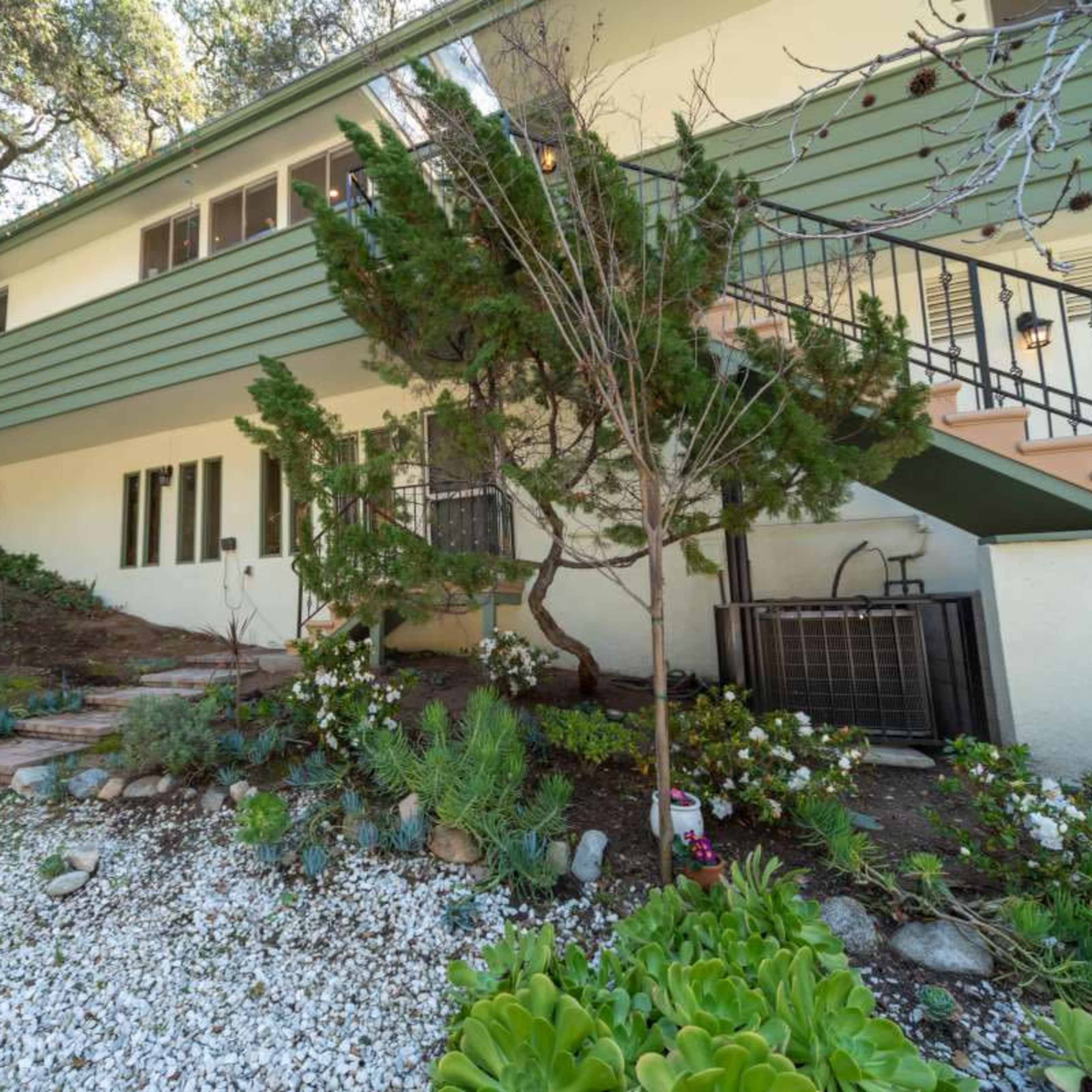 A two-story house with green siding sits on a sloped yard featuring a pathway of stones and various plants.