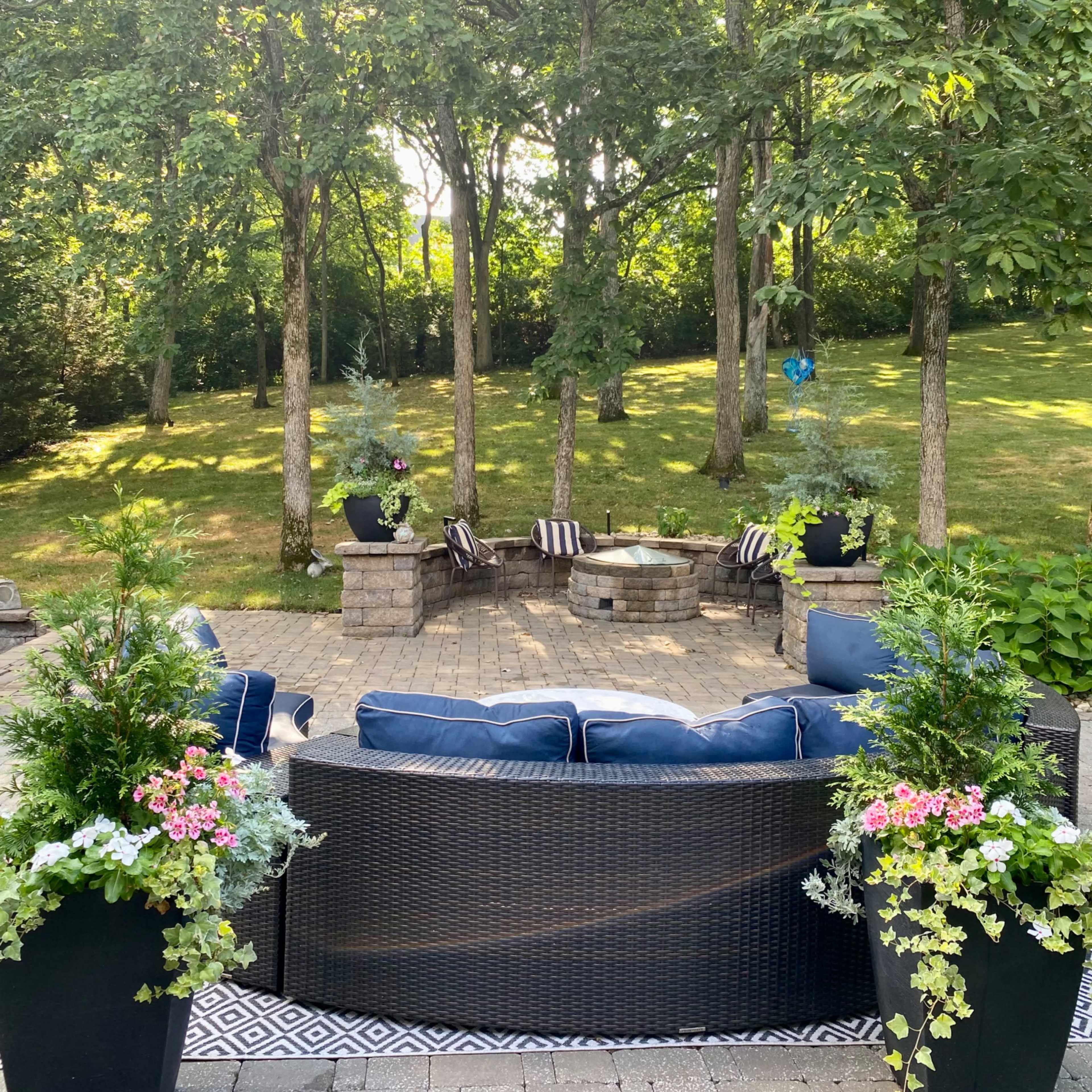 A patio with a seating area surrounded by potted plants and a fire pit is set against a backdrop of trees.