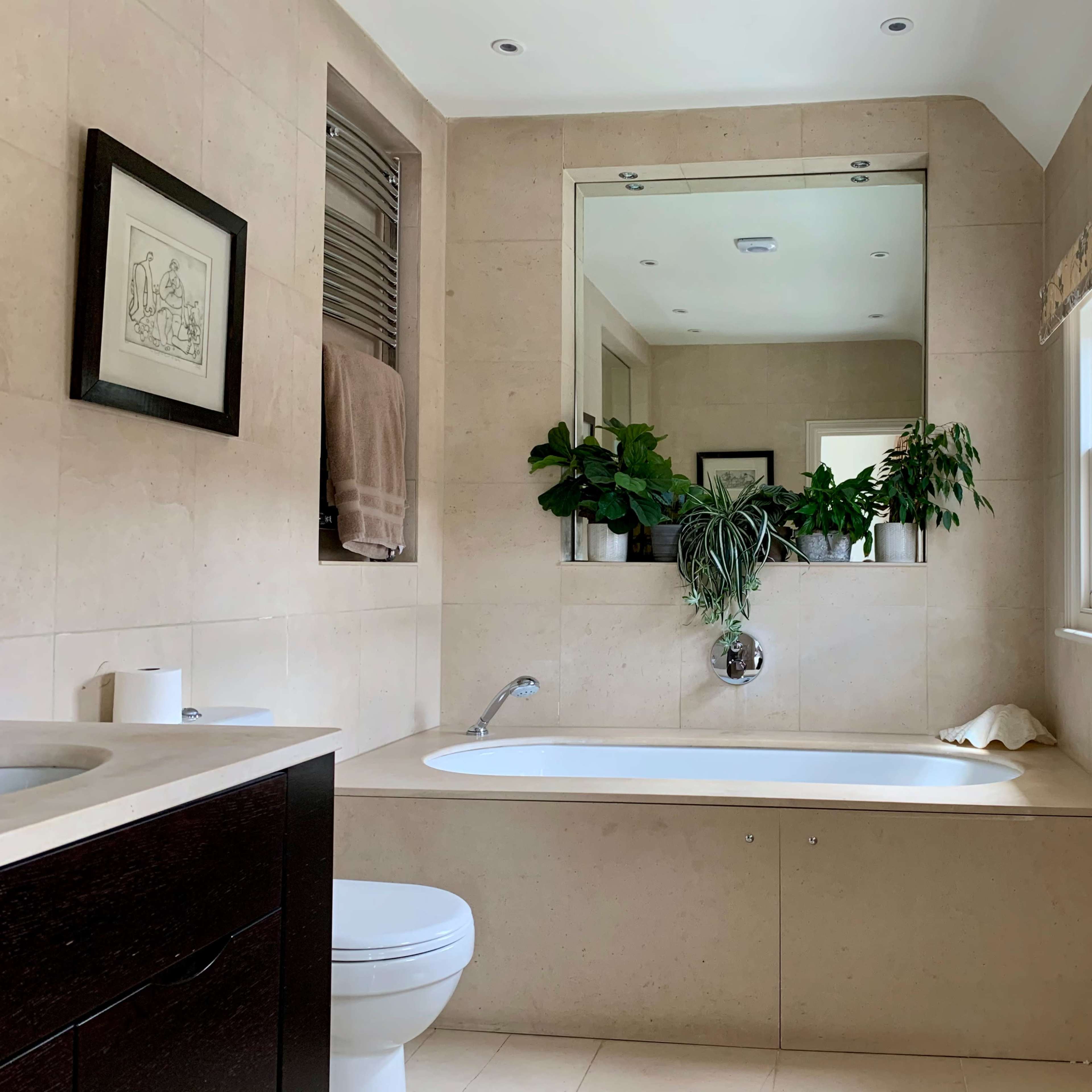 A spacious bathroom featuring a bathtub positioned below a large mirror, surrounded by greenery and light-colored stone walls.