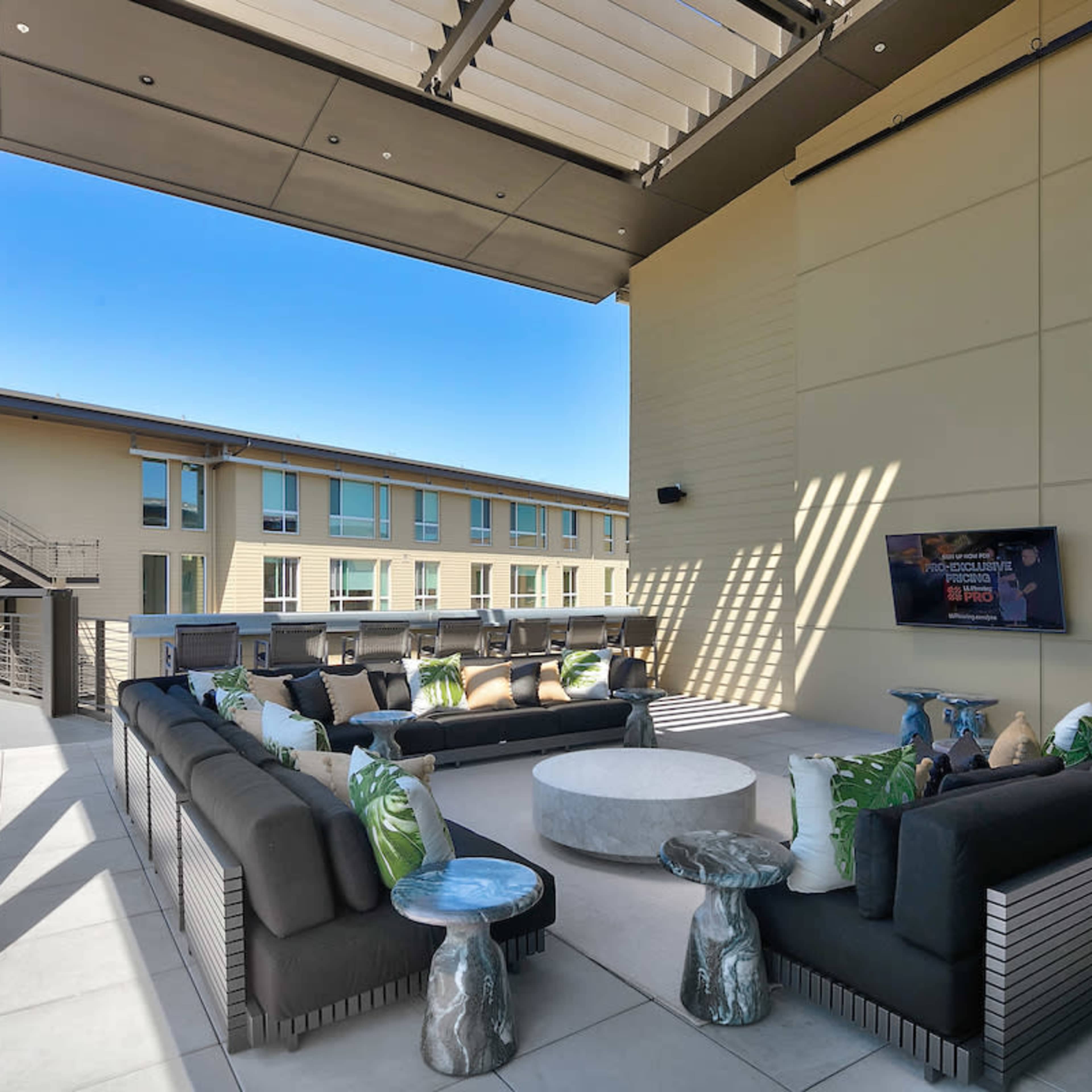 A modern outdoor lounge area with sectional seating, a round table, and a television mounted on the wall, surrounded by a building with multiple windows.