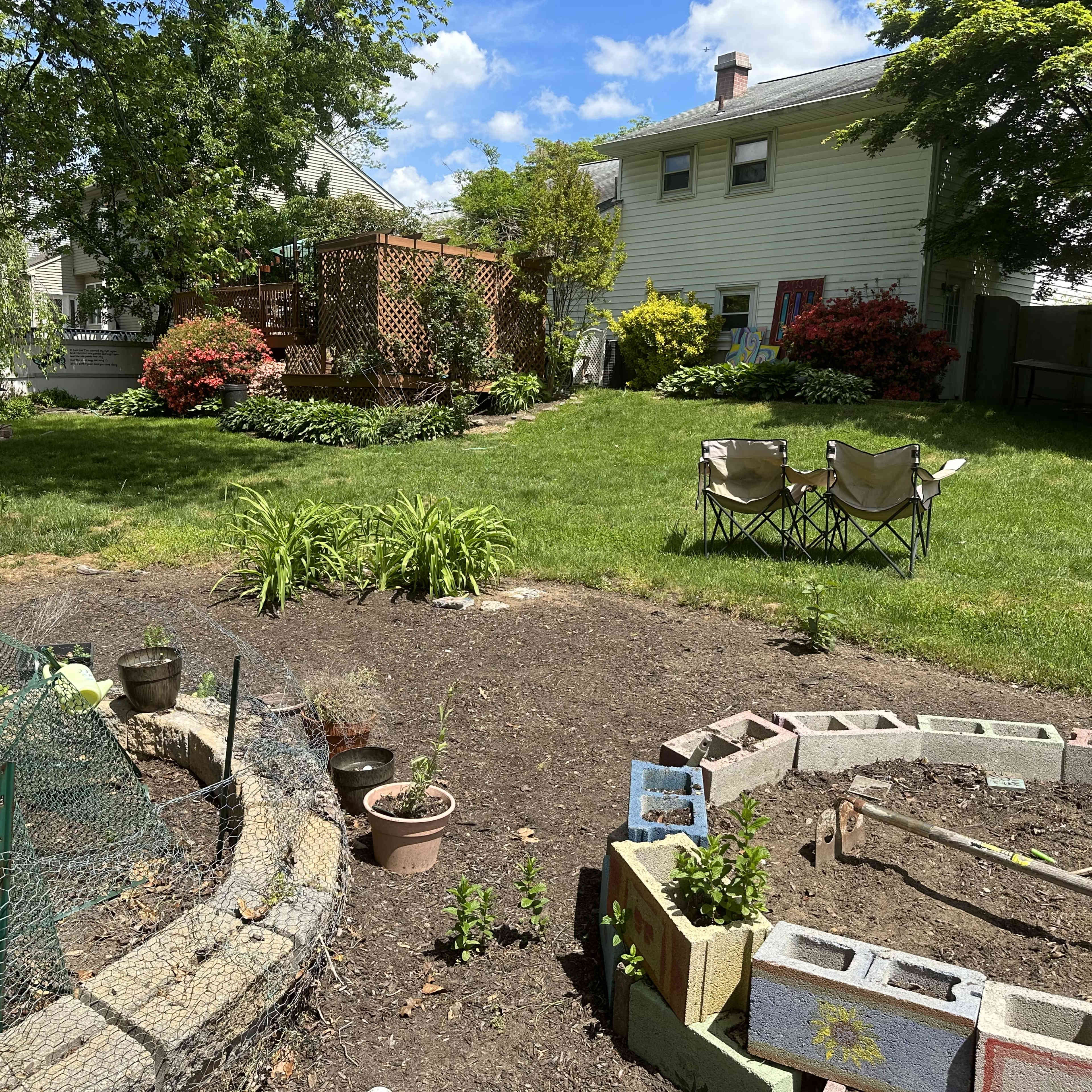 A backyard garden features a circular stone bed with young plants, a fenced area with potted plants, and two chairs facing a lush green lawn and a wooden deck in the background.
