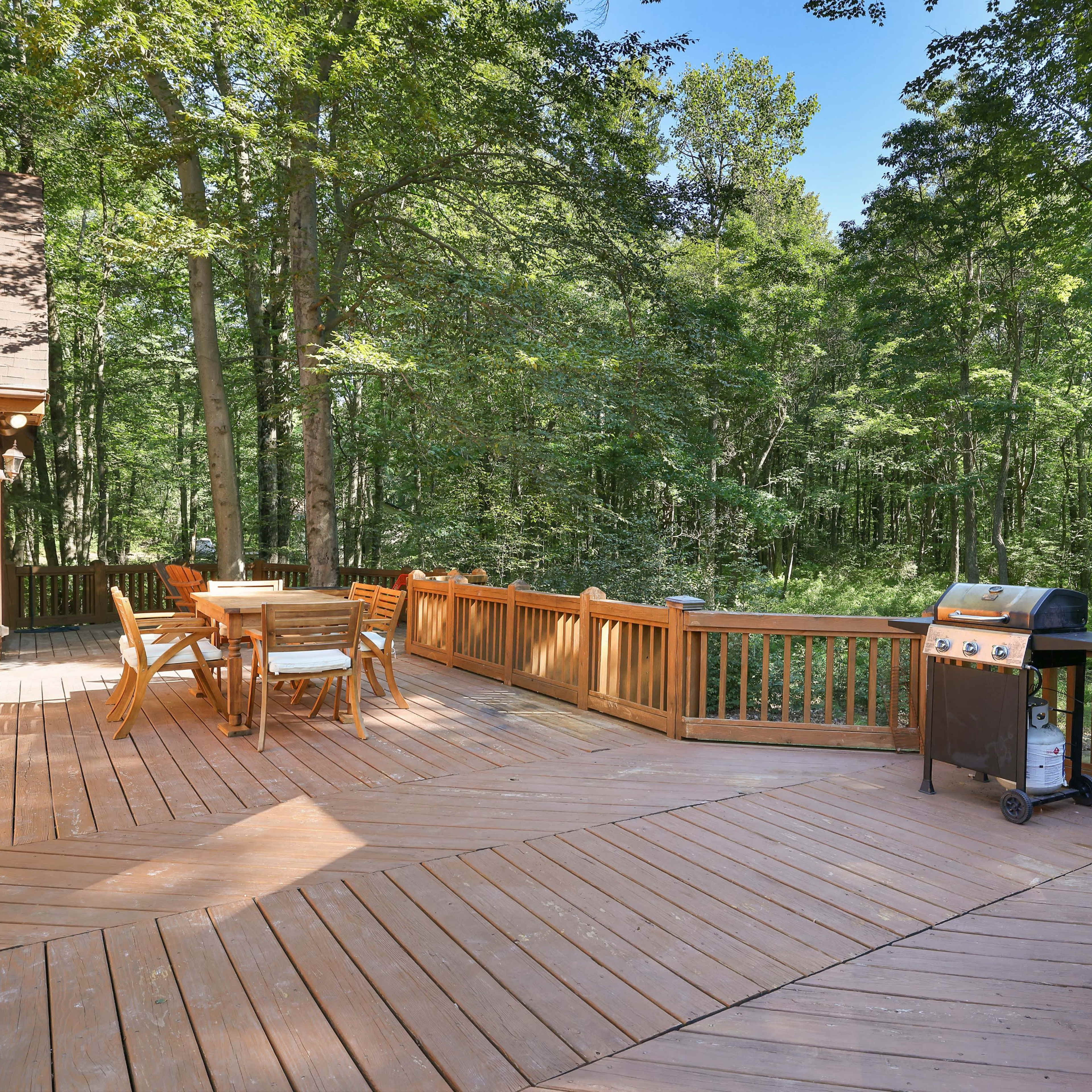 A wooden deck features a grill and several outdoor dining tables surrounded by trees.