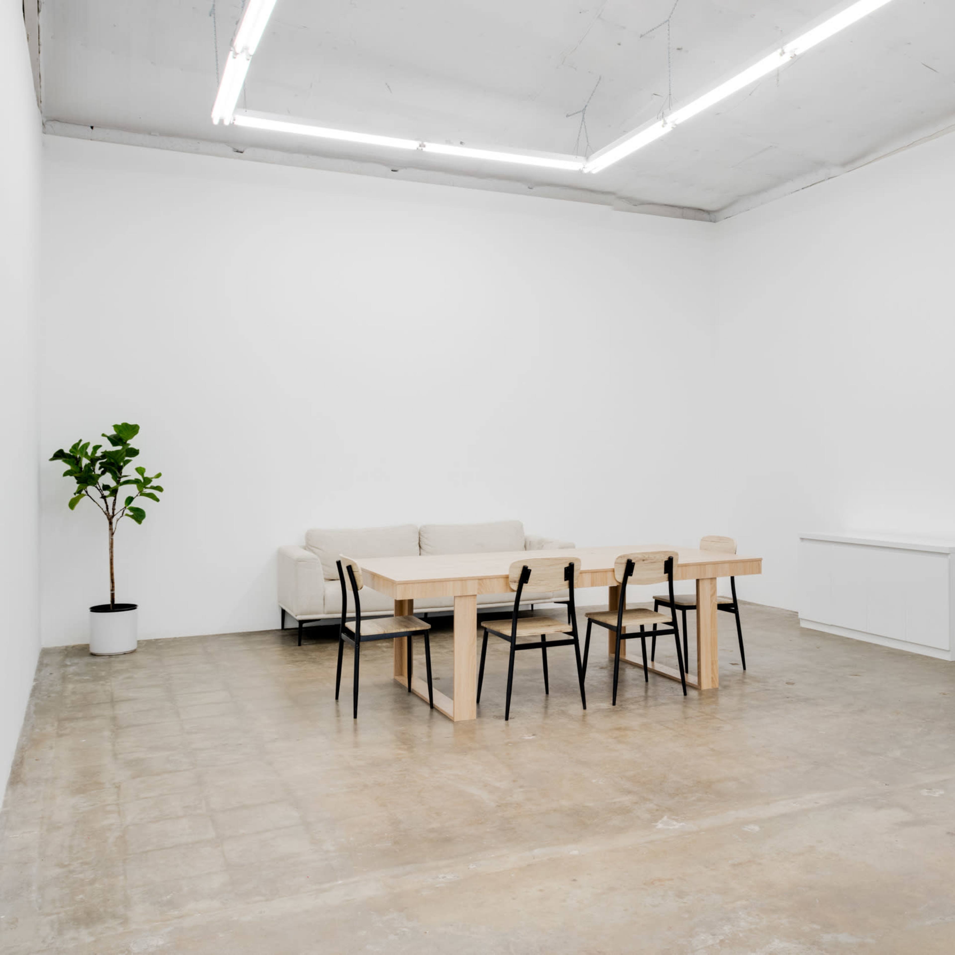 A minimalist indoor space with a wooden dining table surrounded by chairs, a sofa, a potted plant, and a circular mirror above a dark cabinet.