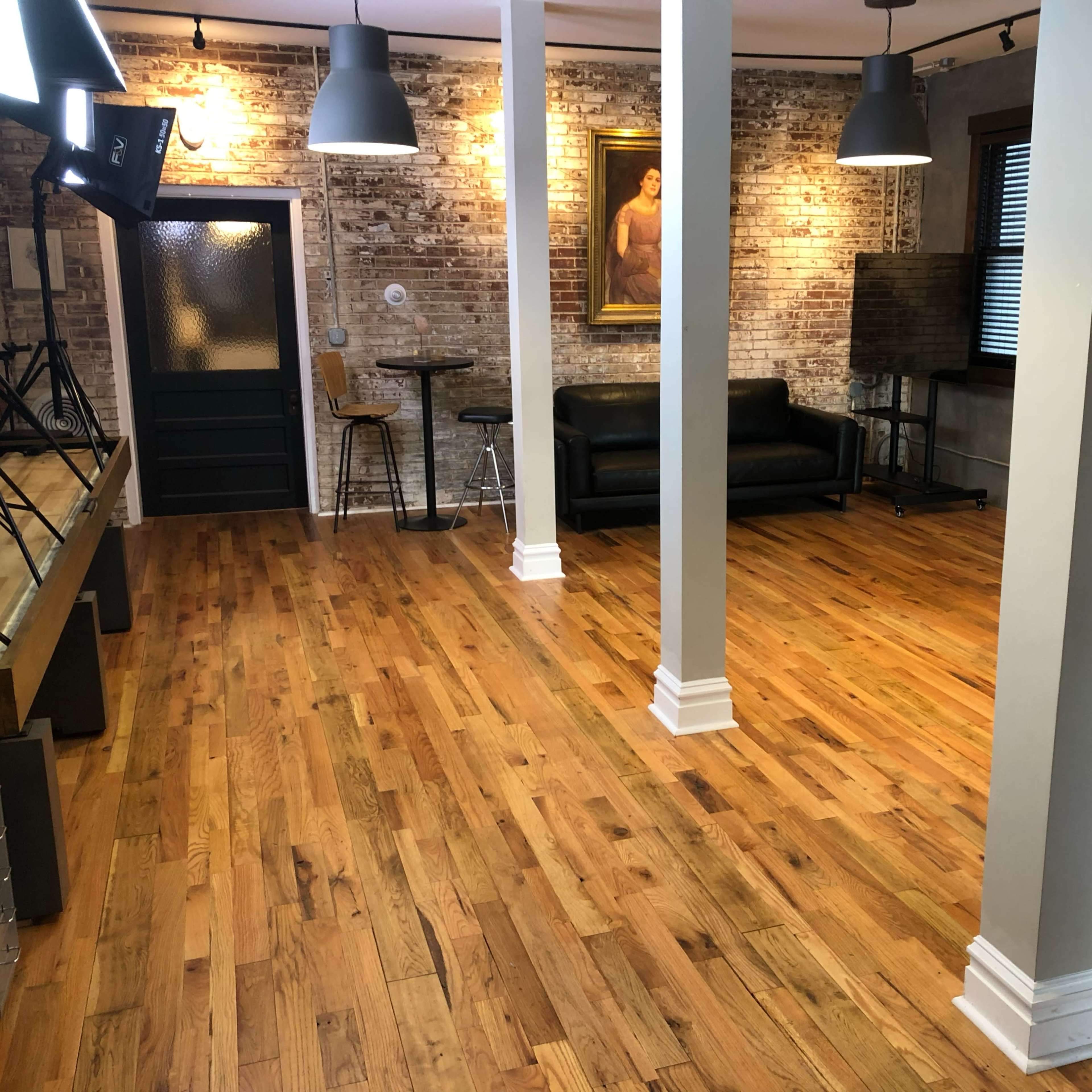 A spacious room with wooden flooring, columns, a dark couch, and a painting on the exposed brick wall.