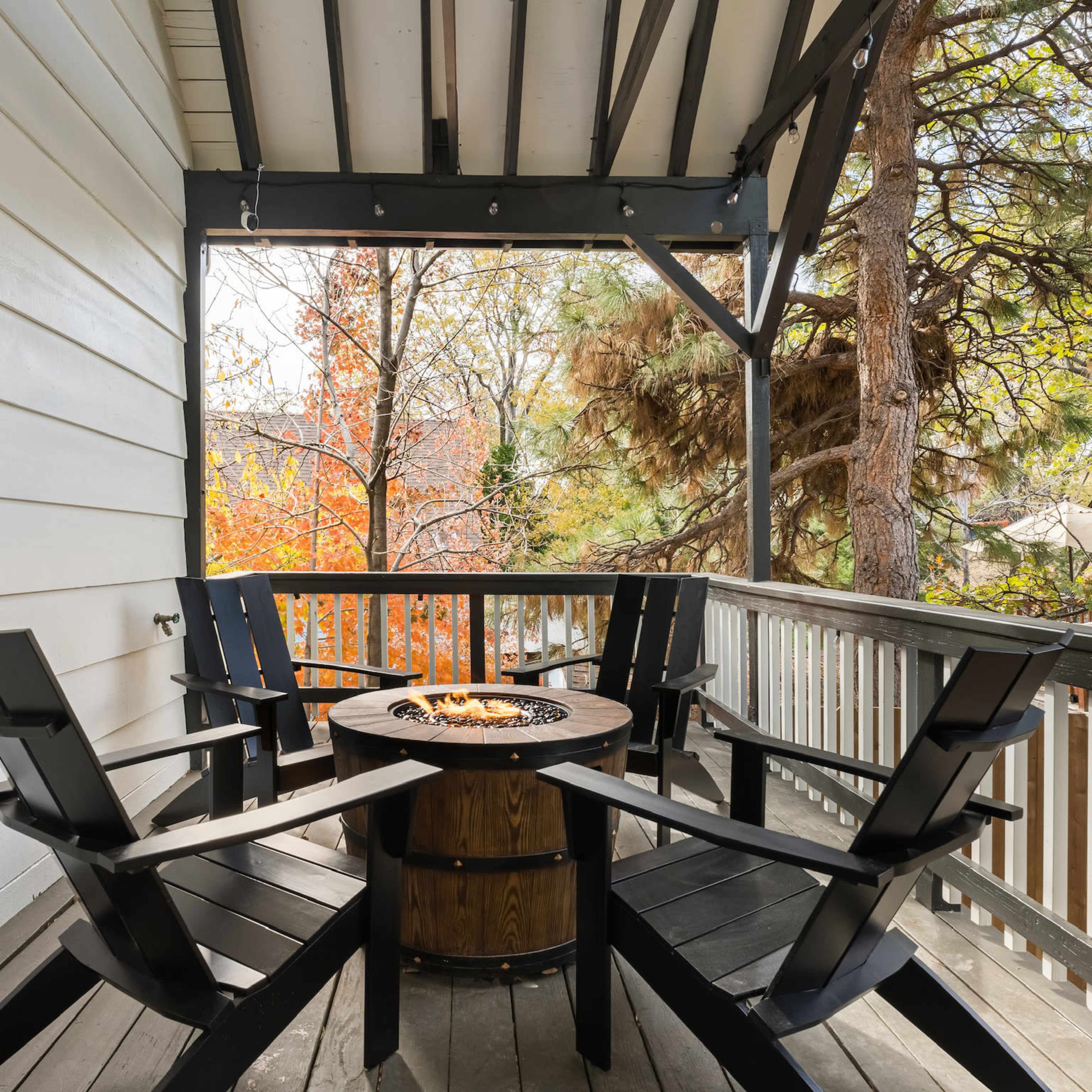 A wooden deck featuring four black adirondack chairs arranged around a circular fire pit.