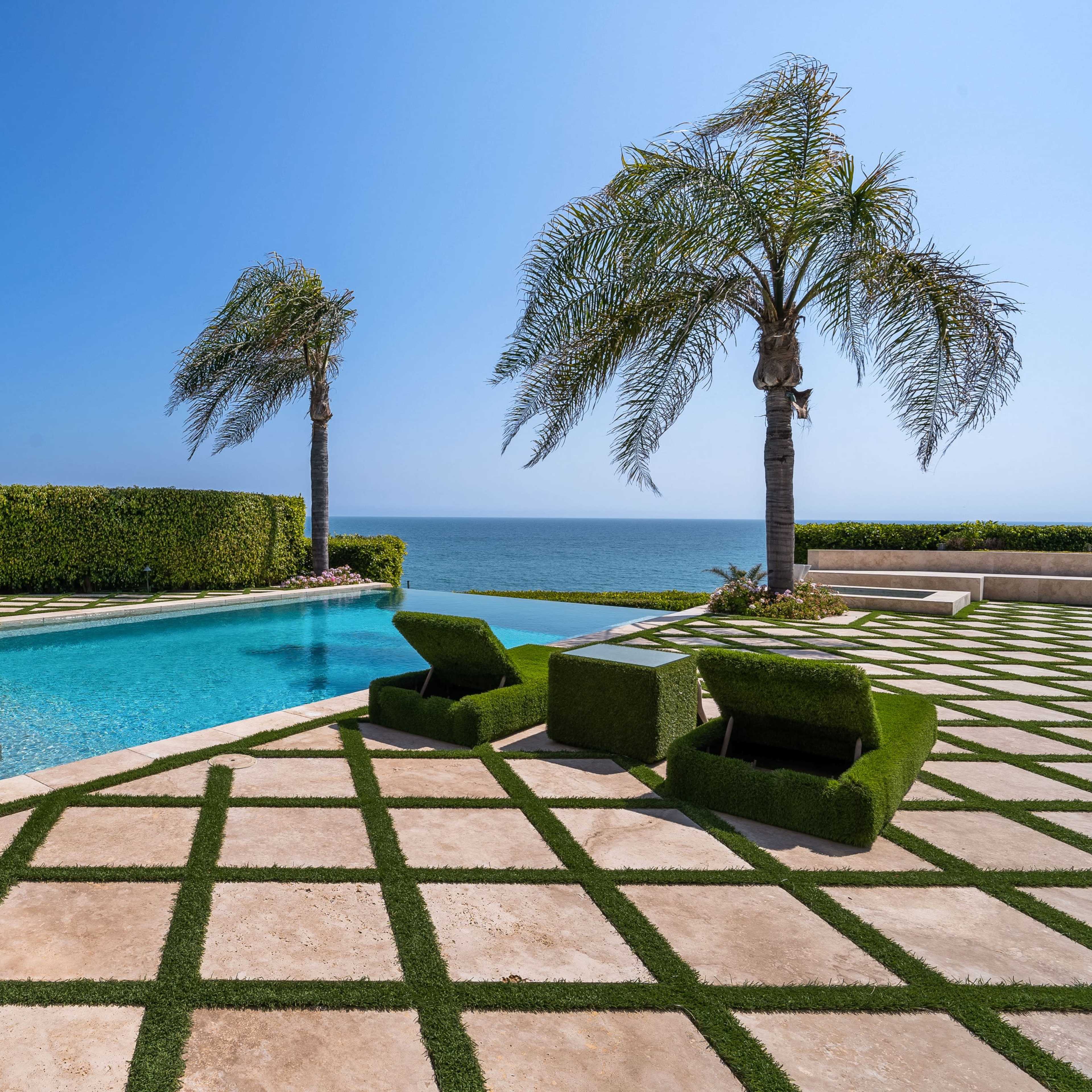 A pool area features green lounge chairs and neatly arranged tiles, surrounded by palm trees and overlooking the ocean.