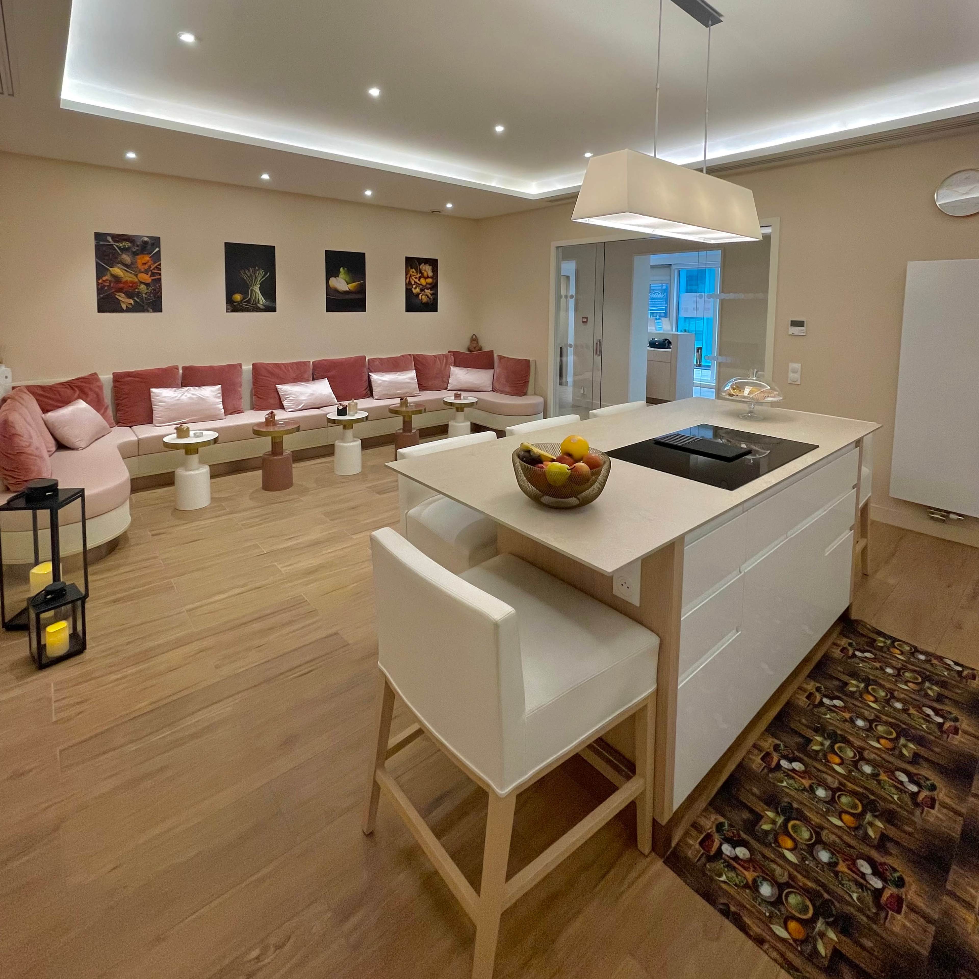 A modern living room features a pink sectional sofa, a sleek kitchen island with a bowl of fruit, and ambient lighting throughout.