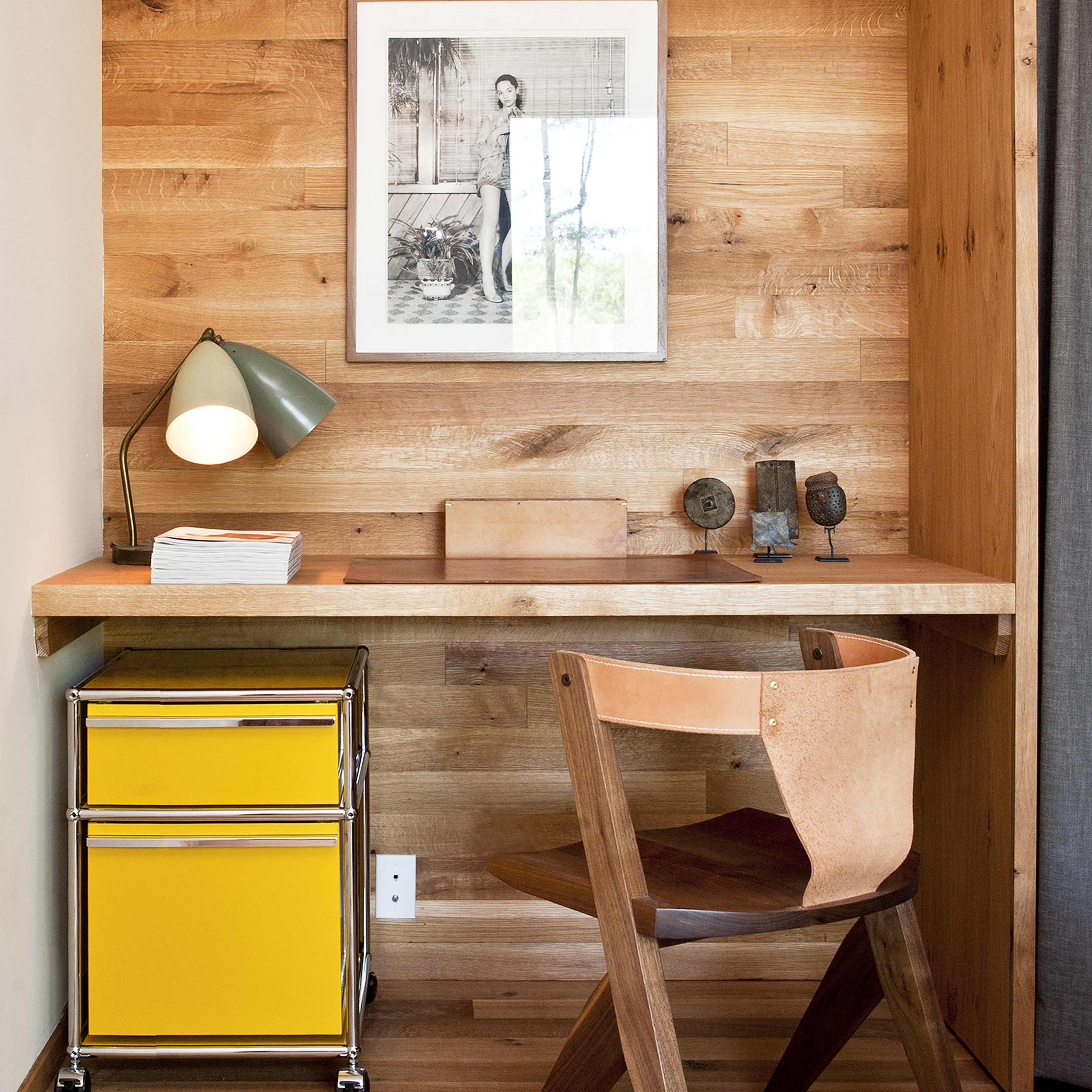 A small workspace featuring a wooden desk against a wall with light-colored wooden panels, accompanied by a yellow storage cart and a wooden chair.