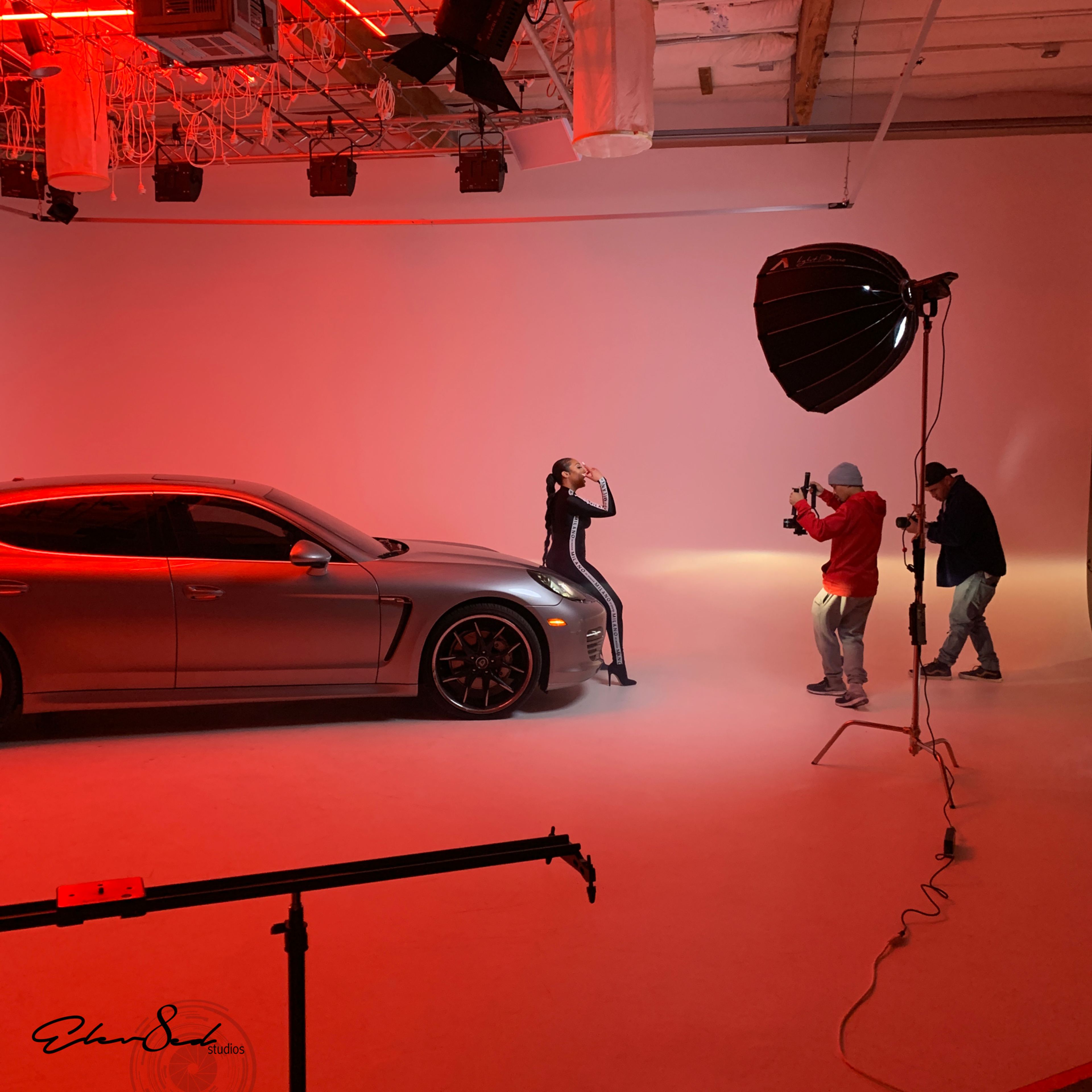 A model poses next to a car while two photographers capture the scene in a studio with red lighting.