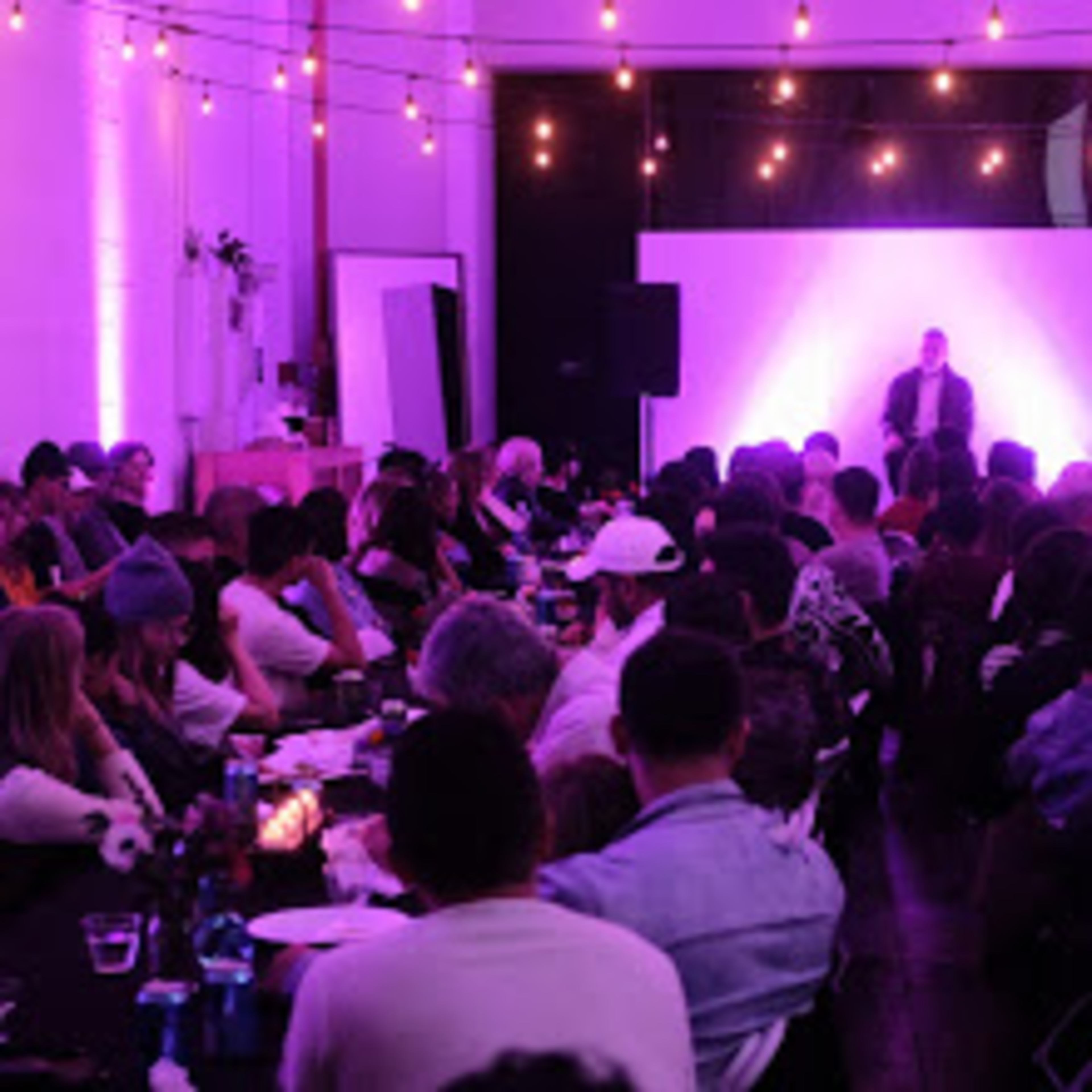 A large audience sits in a dimly lit room with purple lighting, focused on a speaker on stage in front of a blank screen.