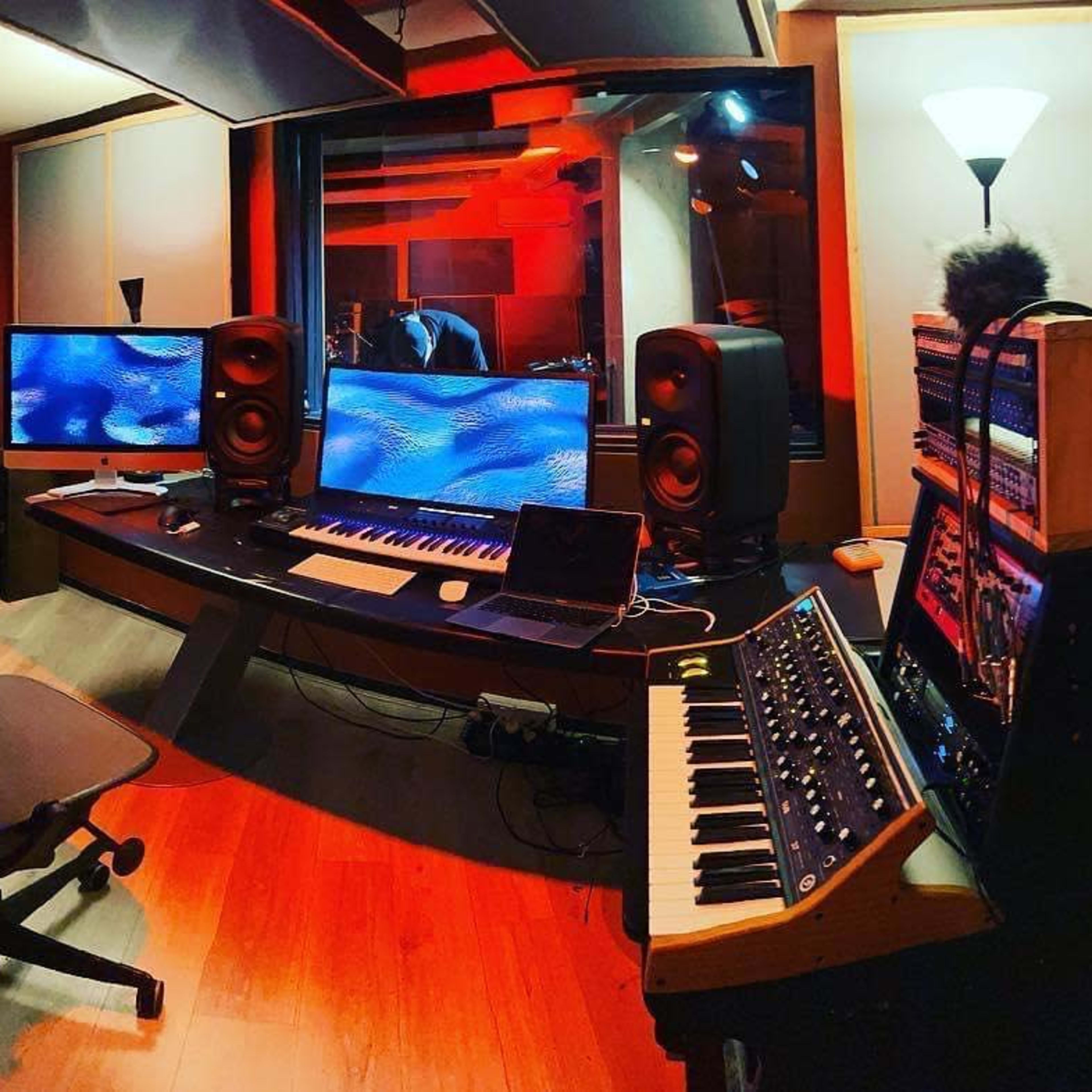 A music studio with electronic instruments, two large monitors, and various audio equipment arranged on a curved desk.