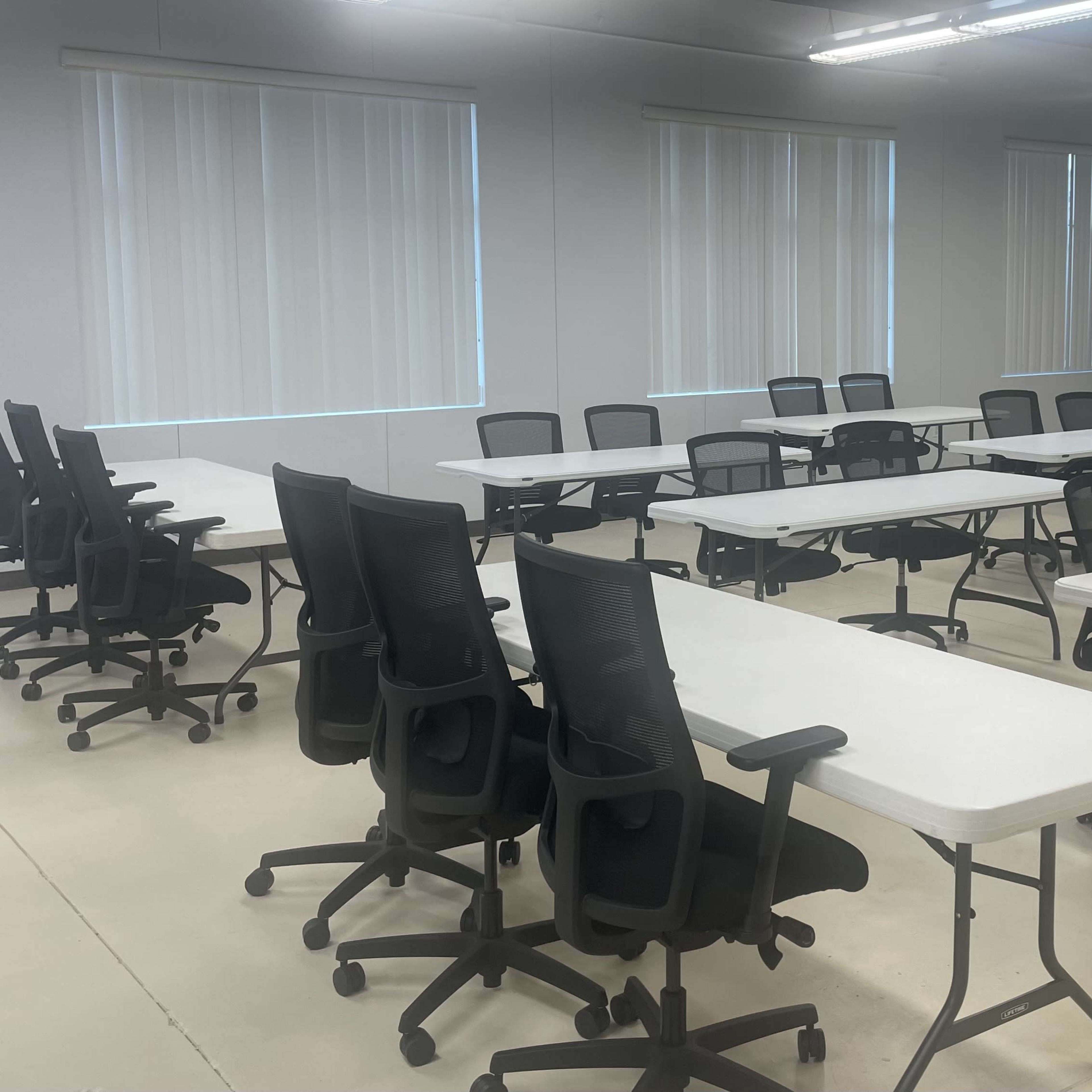 A spacious meeting room features multiple rows of folding tables and black office chairs arranged neatly.