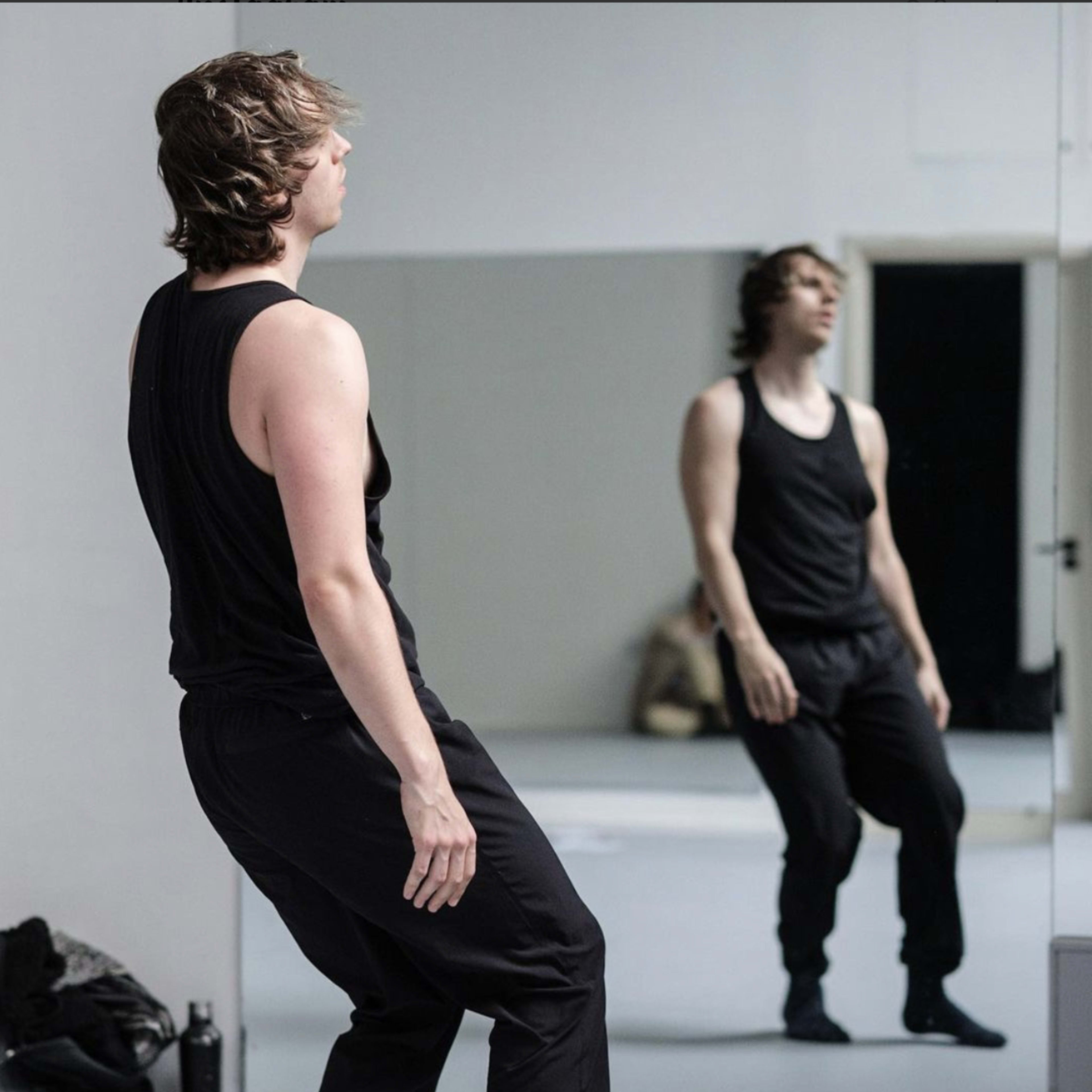 A man in black athletic wear stands in front of a mirror in a dance studio.