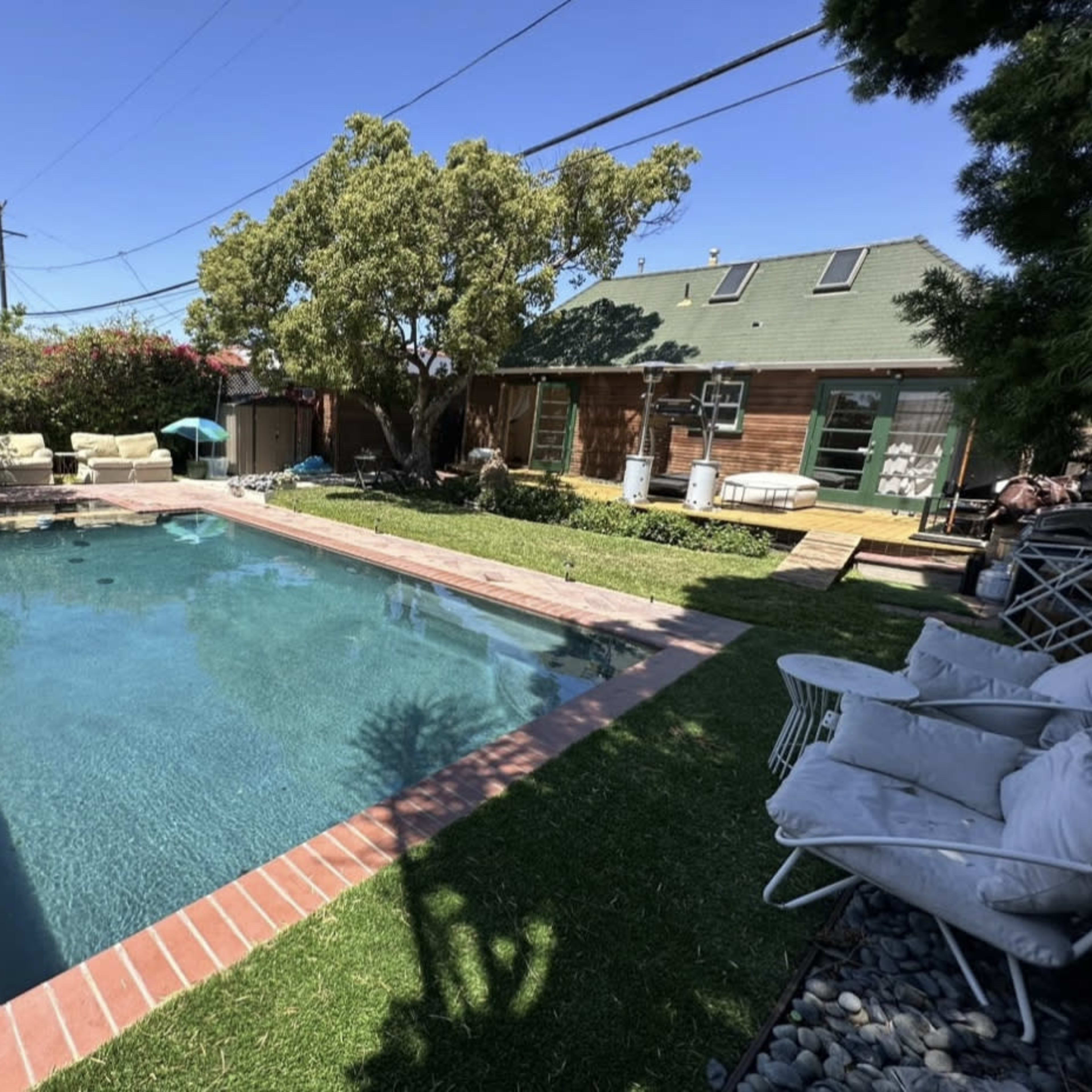 A backyard featuring a rectangular swimming pool surrounded by a brick border, green grass, and some seating areas under trees.