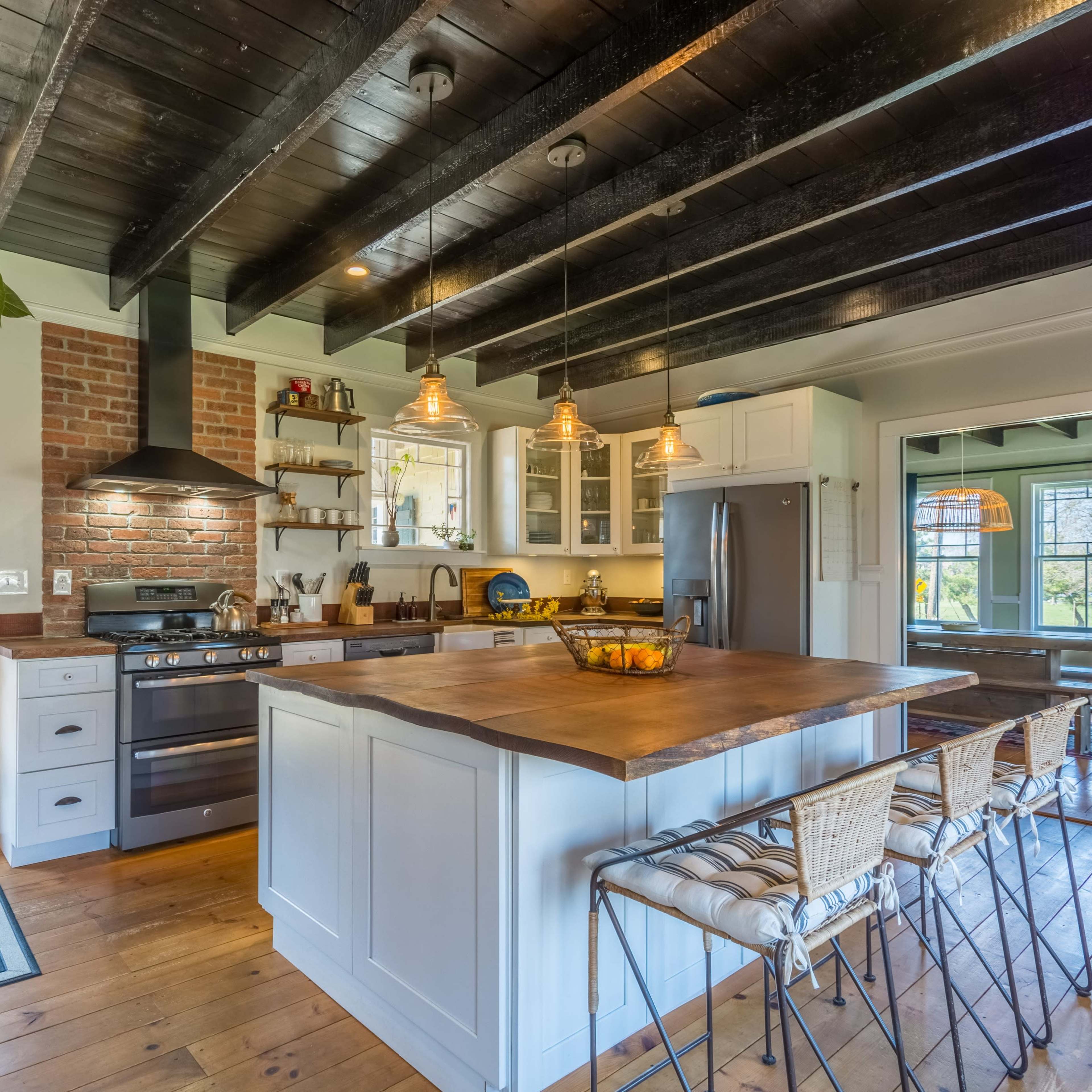 A modern kitchen with a large central island, wooden ceiling beams, and a combination of brick and white cabinetry.