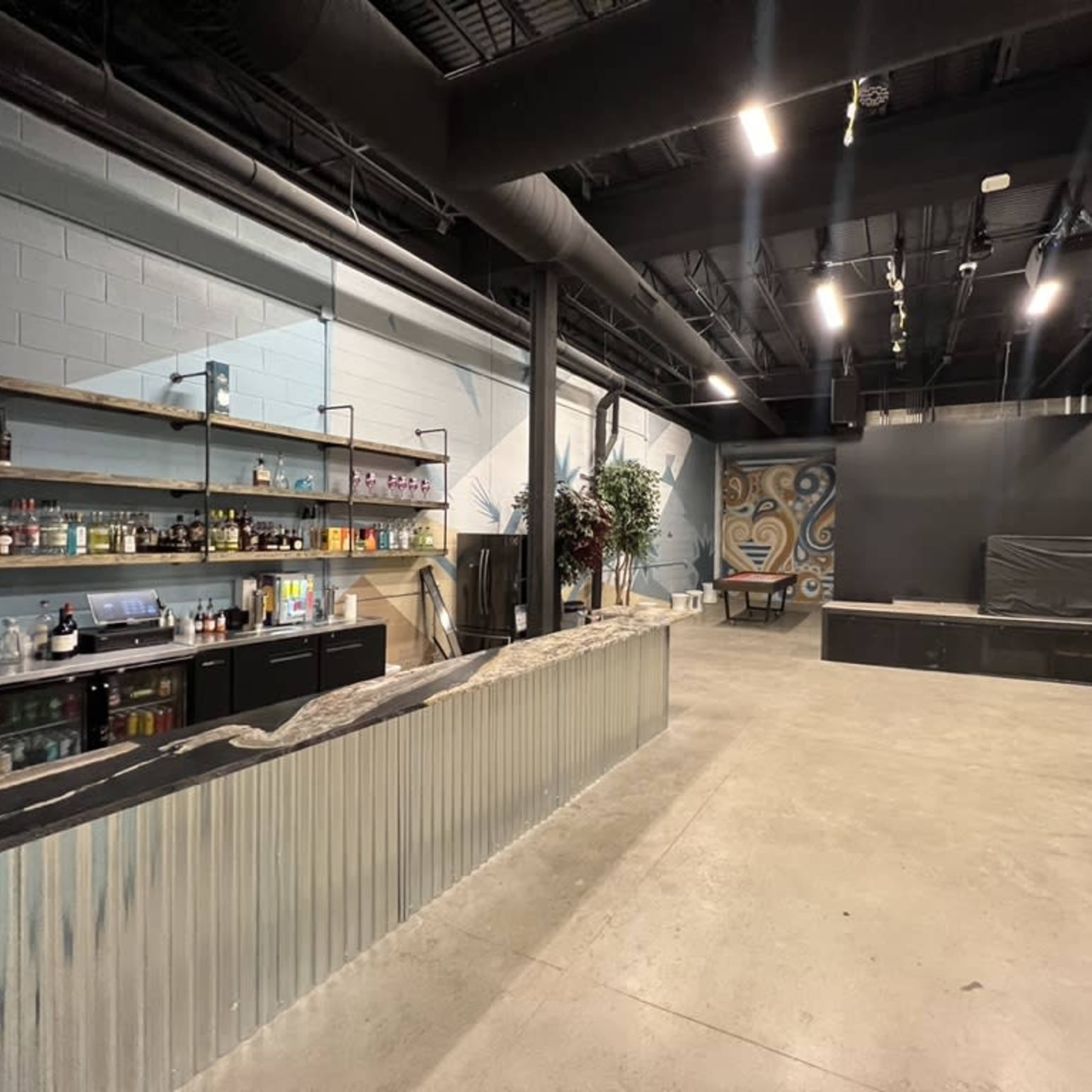 An empty event space with a bar area, shelves lined with bottles, and a large blank stage against one wall.