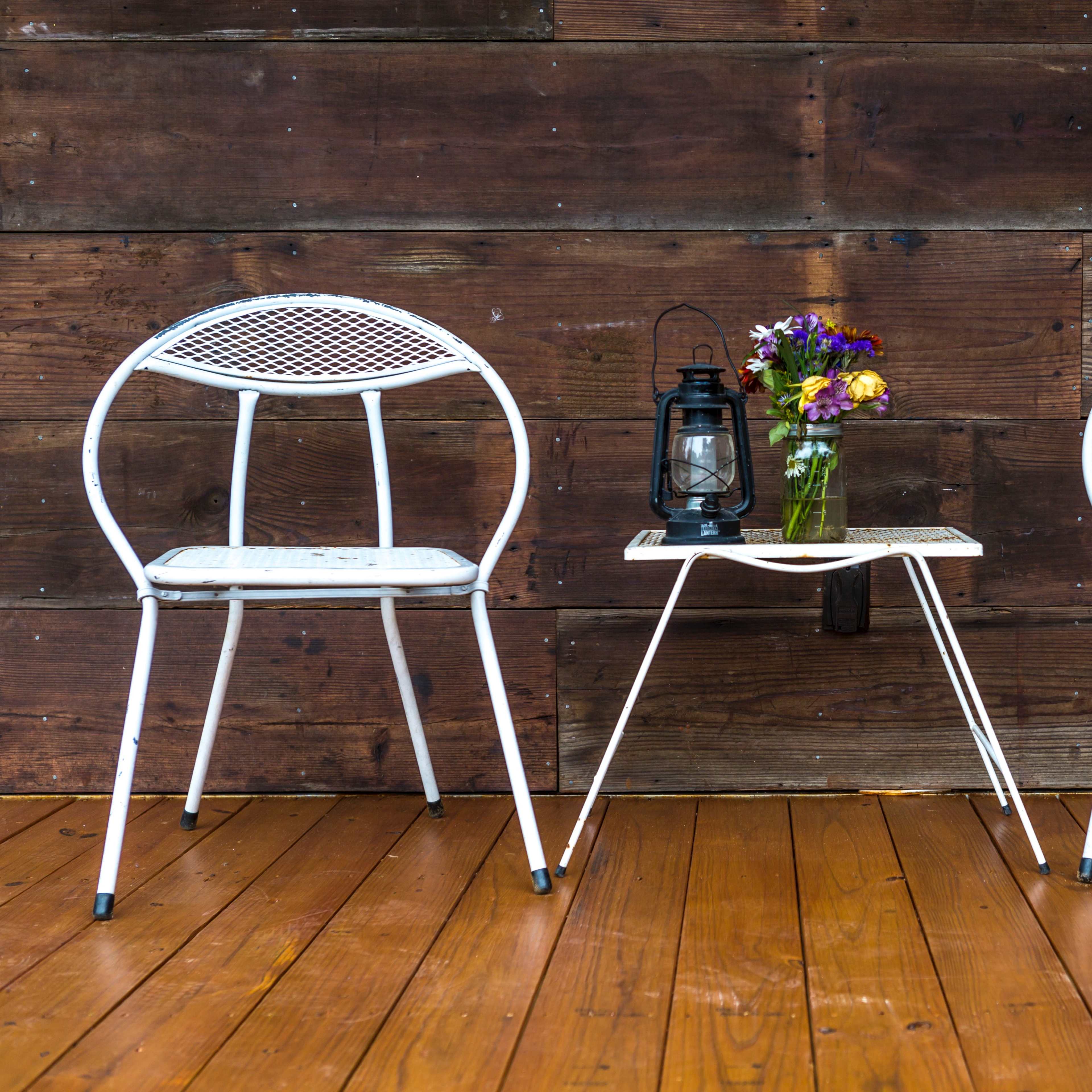 Two white metal chairs and a small table with a lantern and a vase of flowers are positioned on a wooden deck against a rustic wooden backdrop.