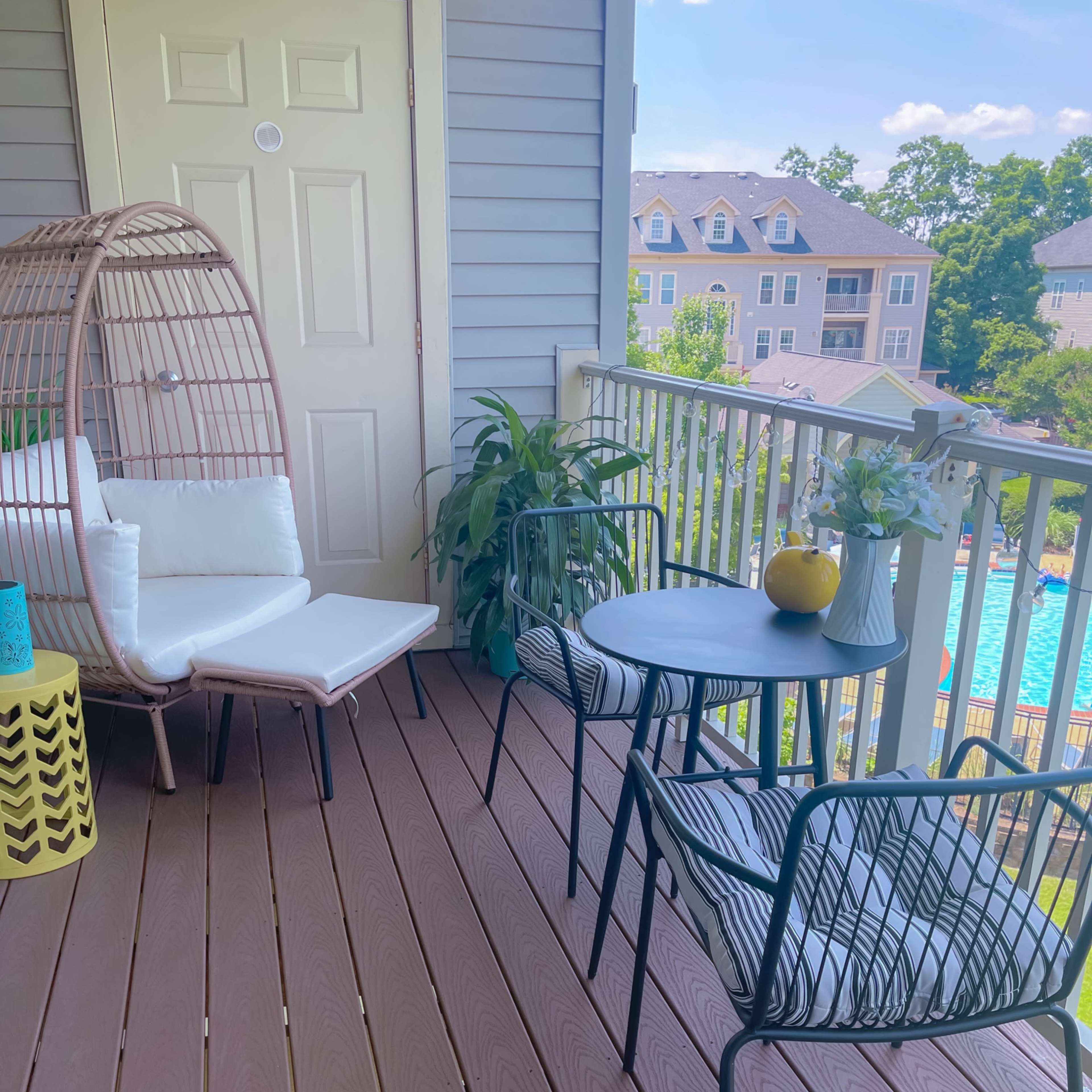 A balcony with a hanging chair, a small table with two chairs, and a view of a swimming pool in the background.