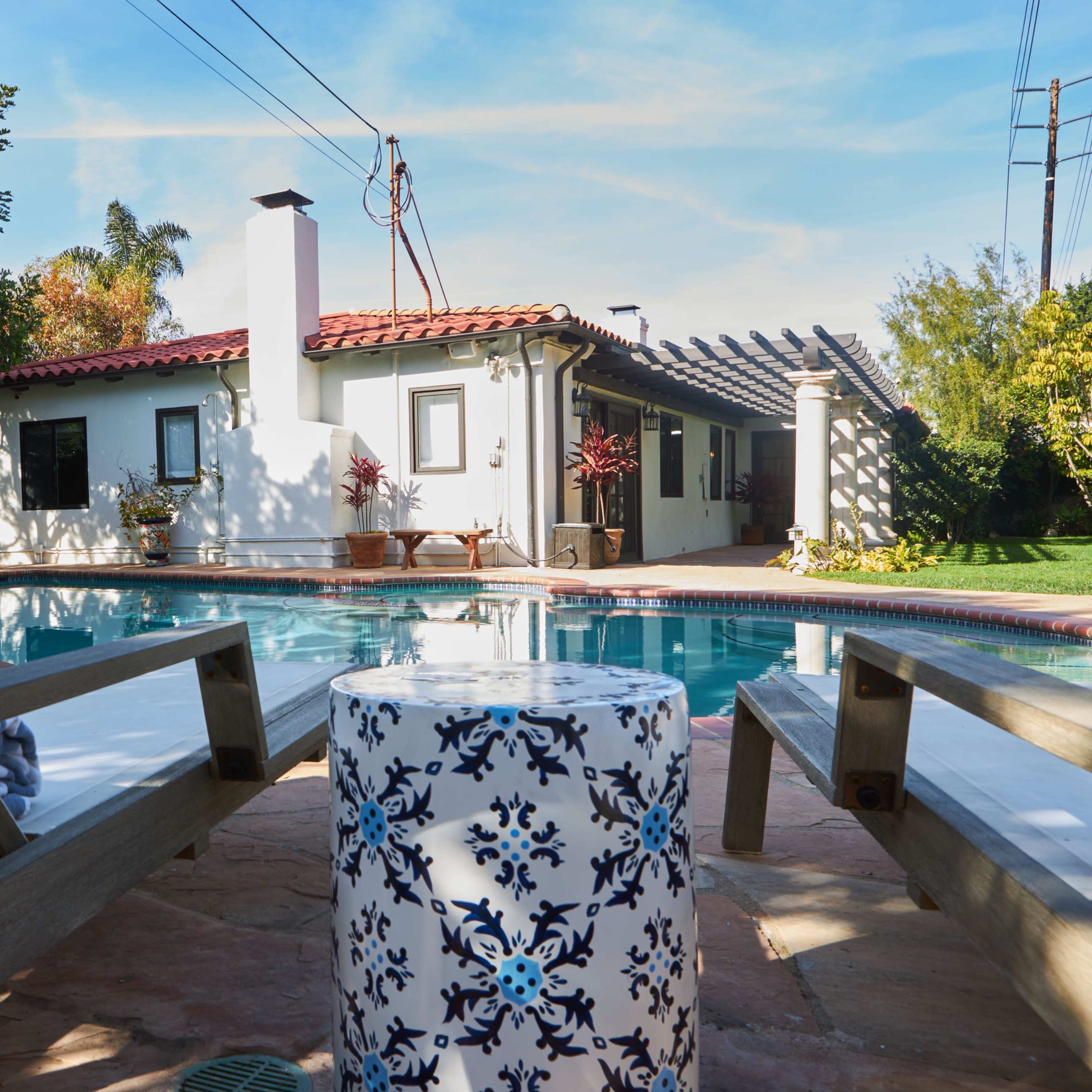 Two wooden lounge chairs flanking a decorative table by a swimming pool, with a house and lush greenery in the background.