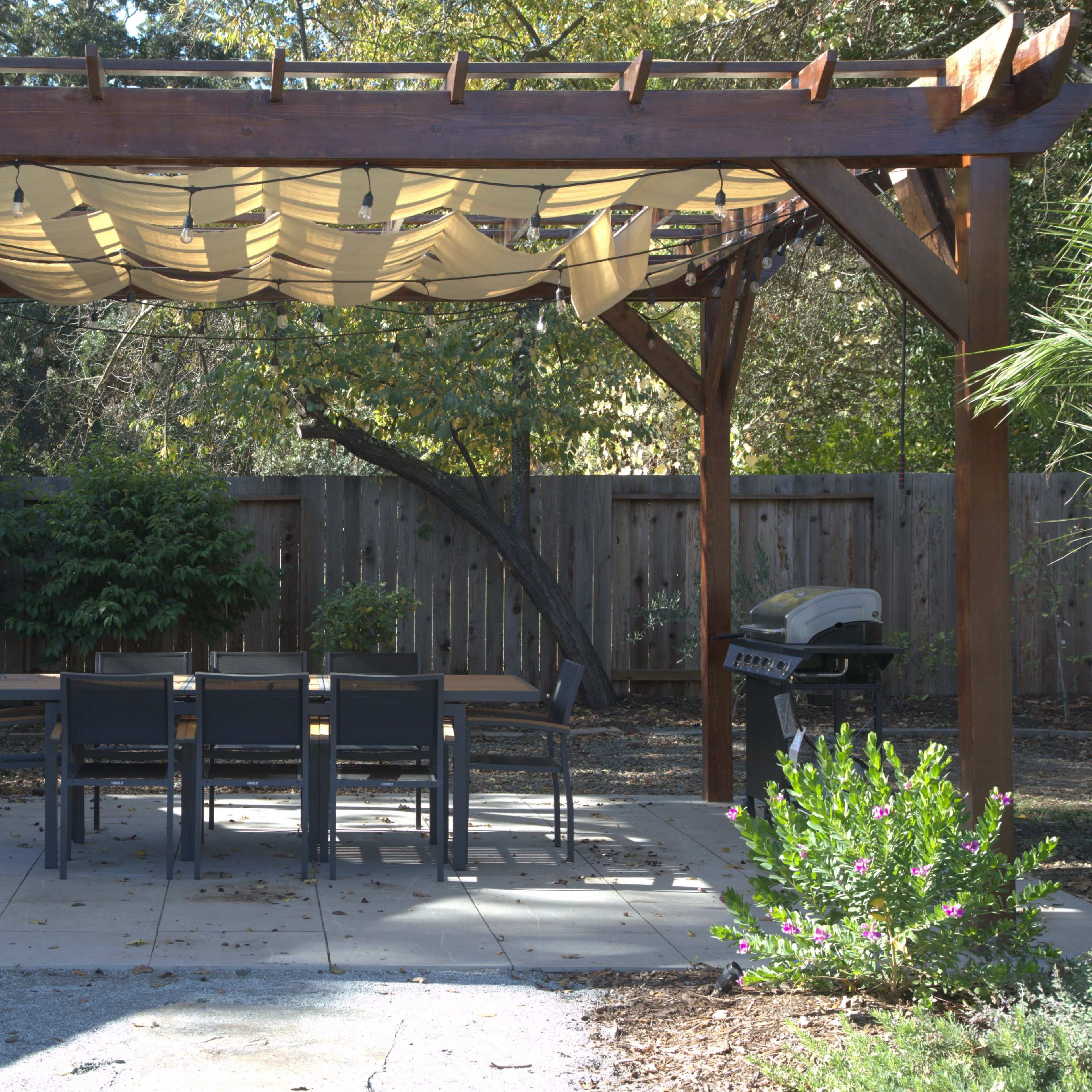 A wooden pergola with a fabric canopy covers a dining table and chairs, situated in a backyard with landscaped borders and a grill nearby.