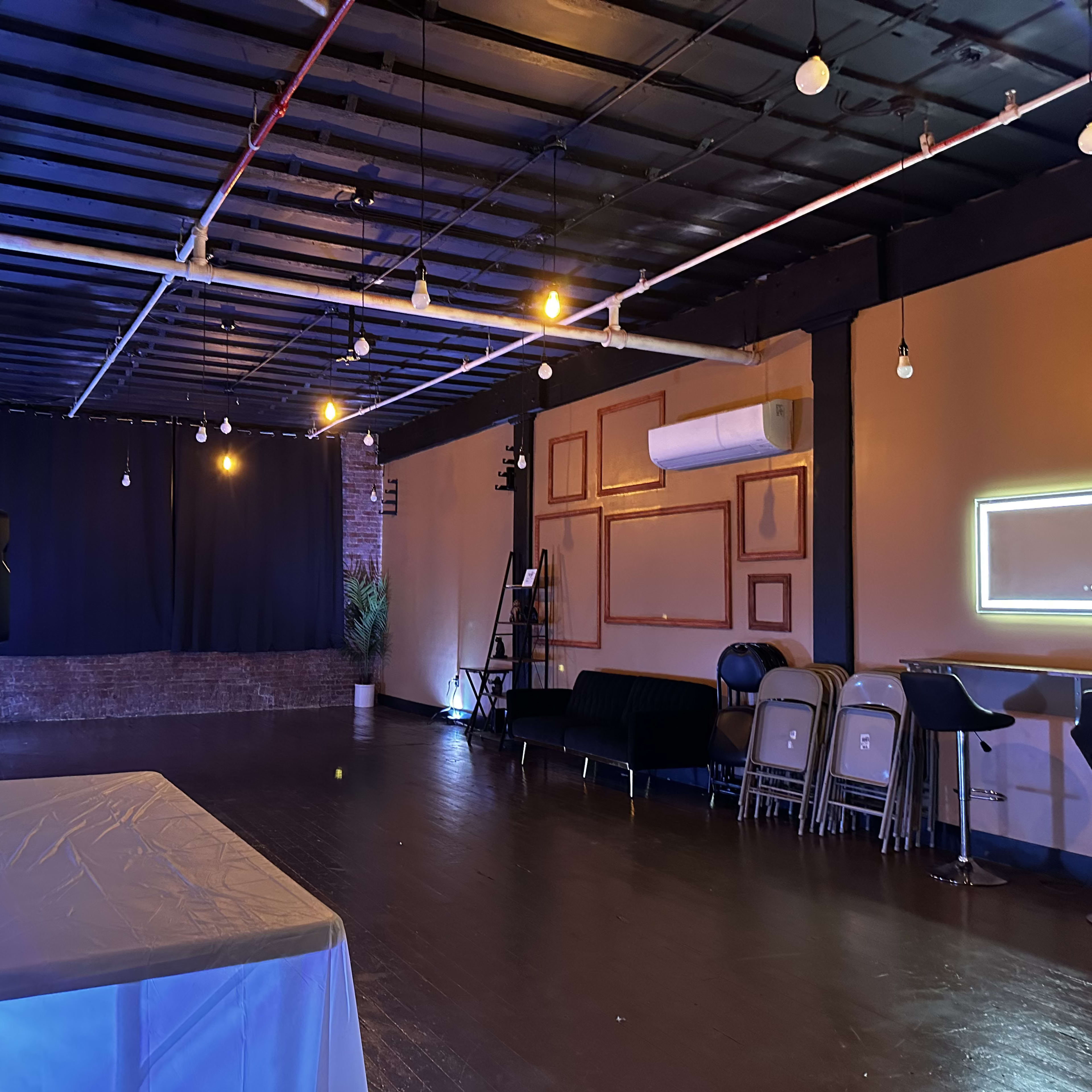 The interior of a dimly lit event space features a black stage area, a white table in the foreground, and rows of metal chairs along the sides.