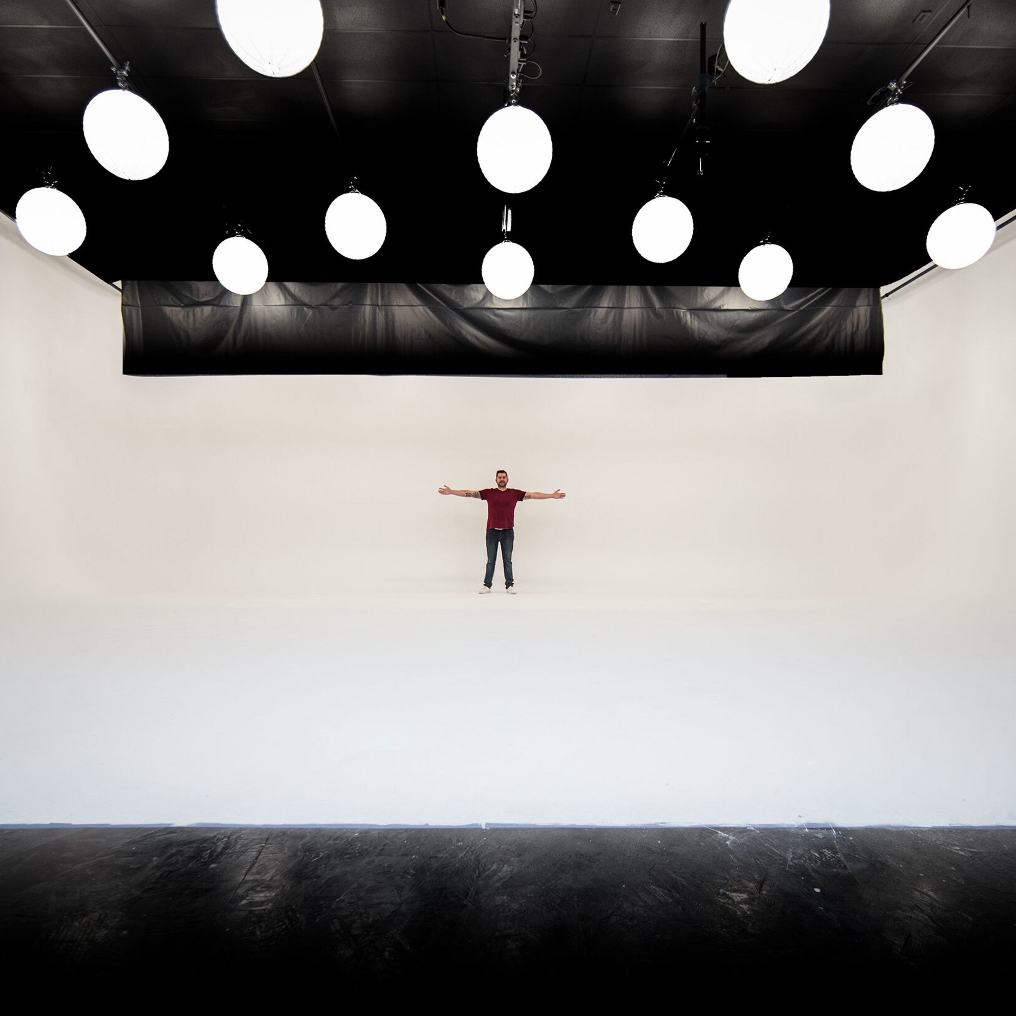 A person stands with arms outstretched in a large, empty studio with white walls and round hanging lights.