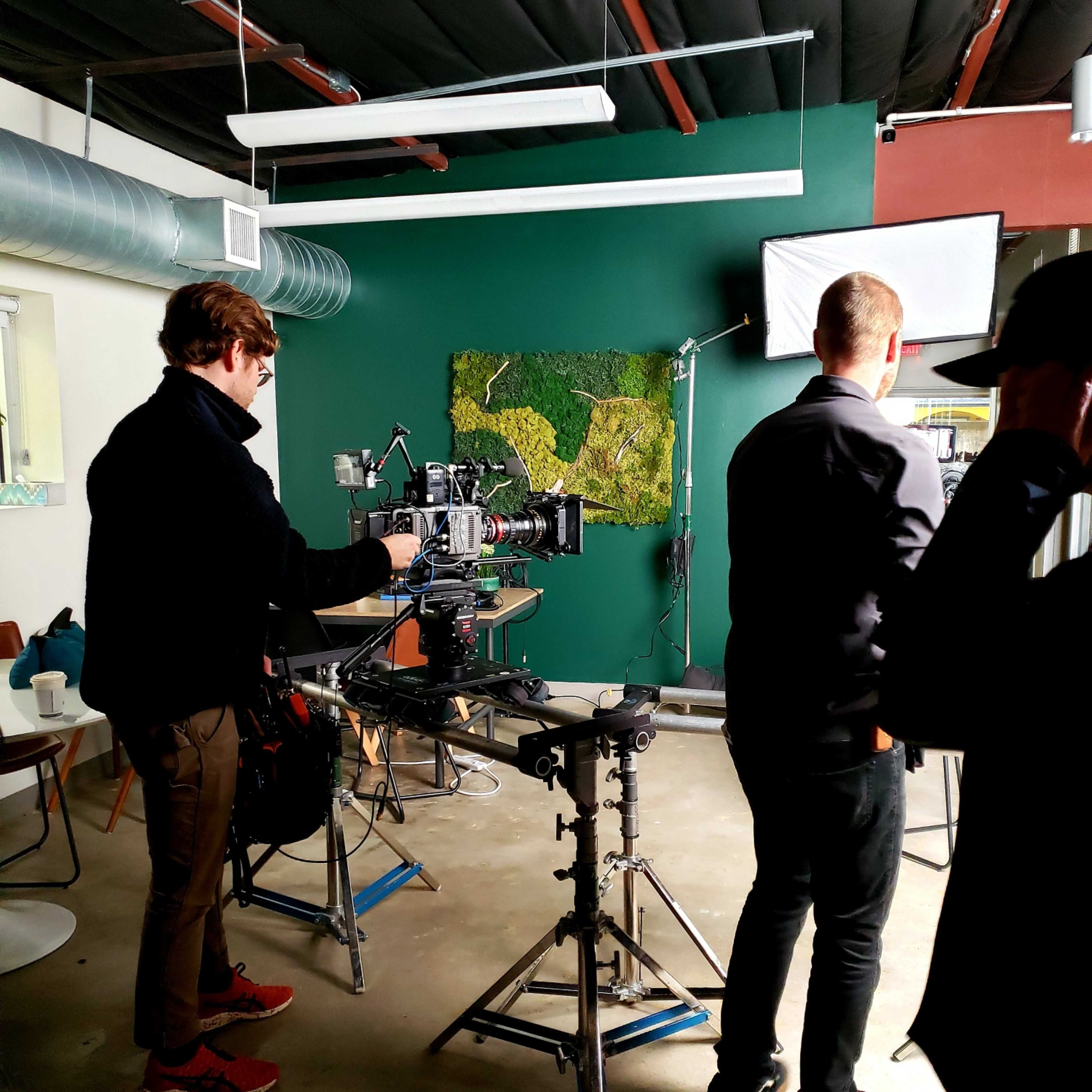 A film crew sets up equipment in an indoor studio space, featuring a camera on a tripod and various lighting rigs.