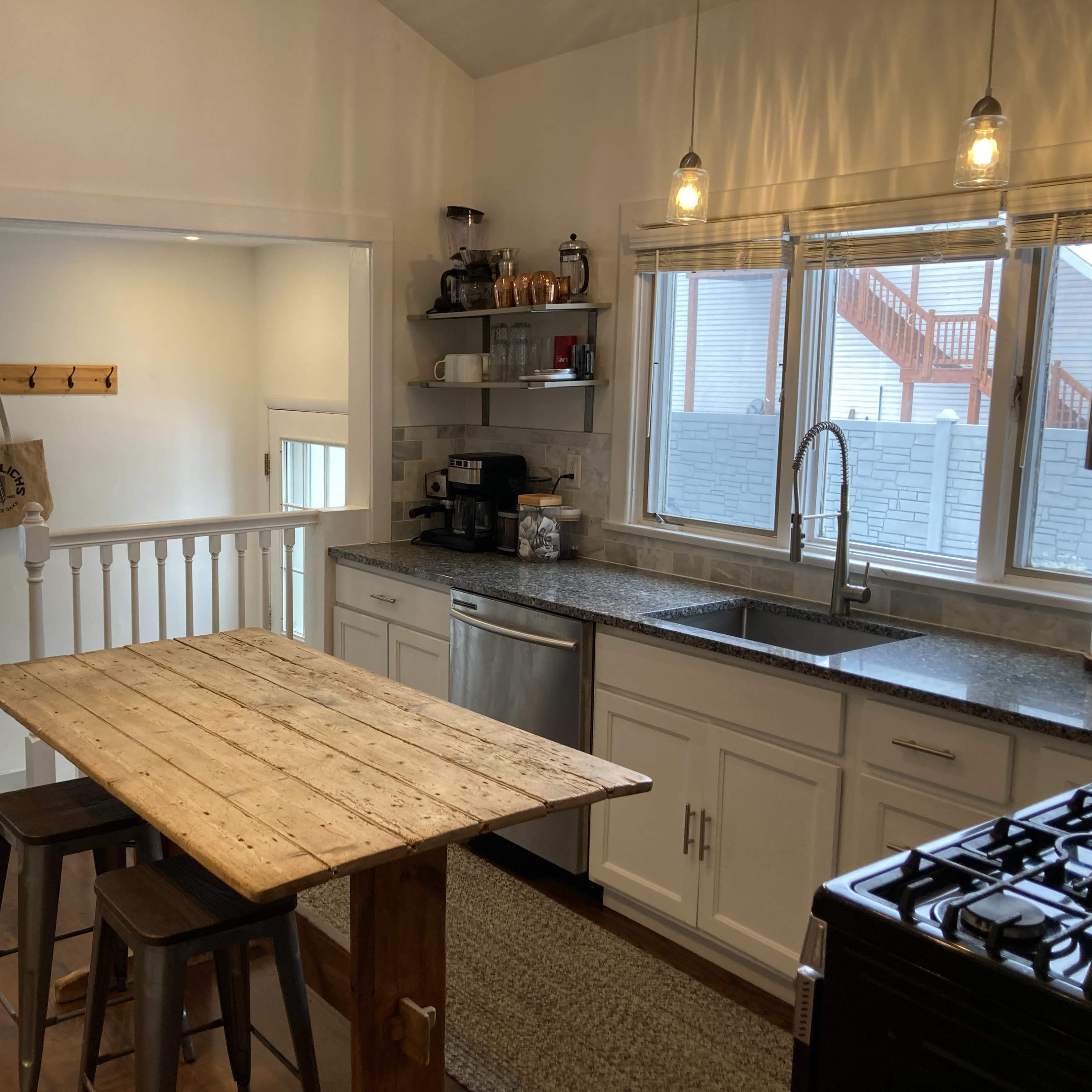 The kitchen features a wooden table with two stools, stainless steel appliances, and a door leading to an outdoor area.