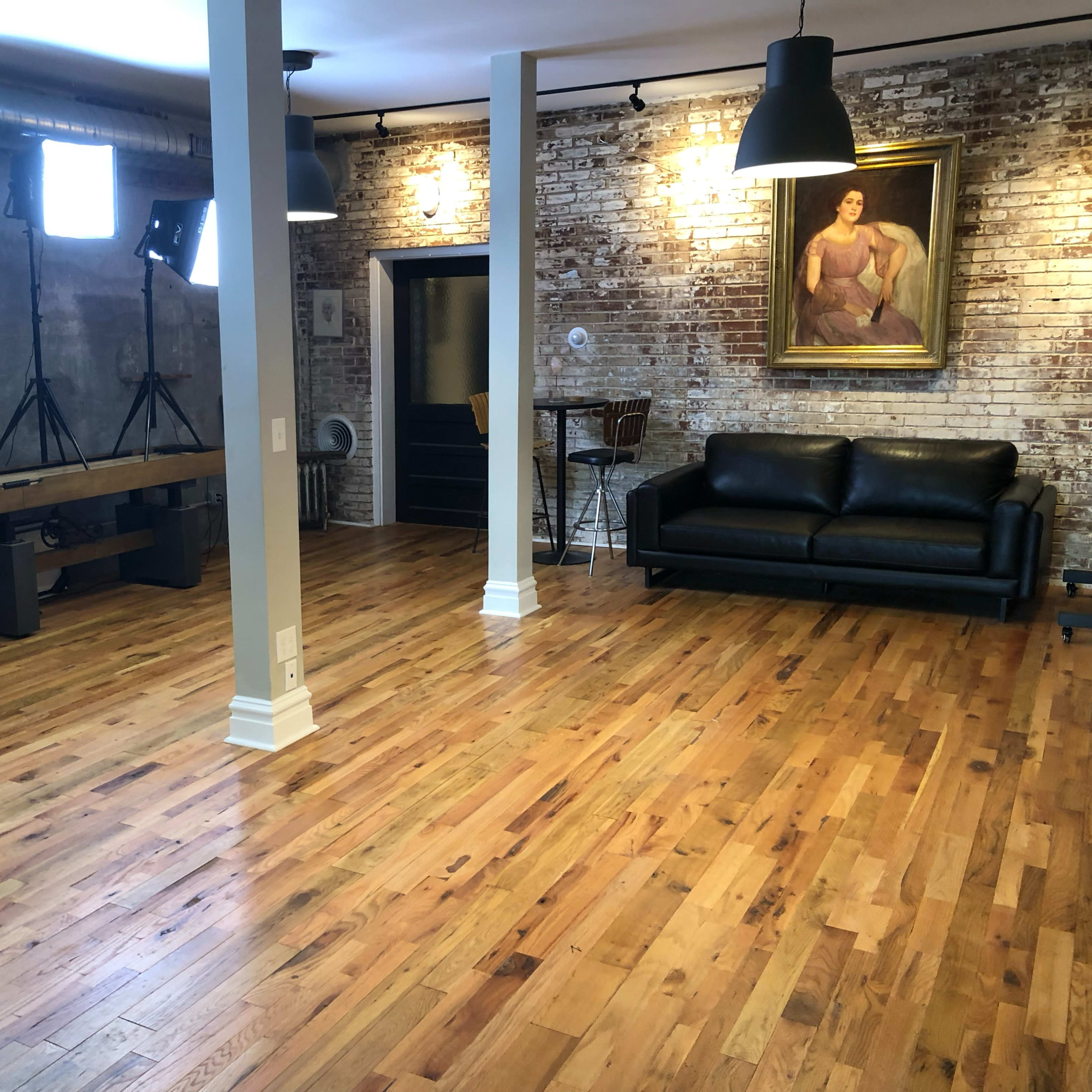 A spacious interior with hardwood flooring, exposed brick walls, a black sofa, and several lighting setups on tripods.