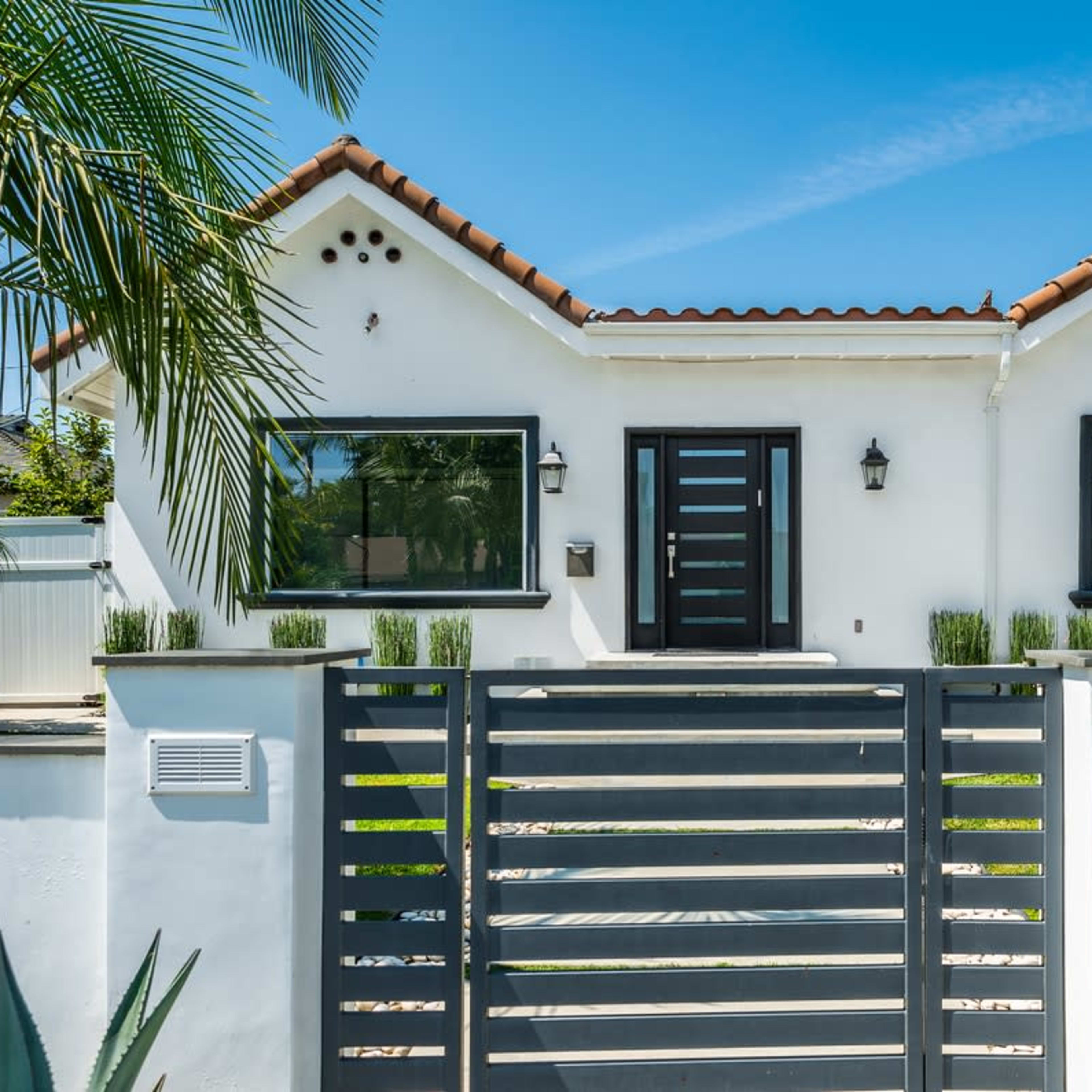 A modern white house with a red-tiled roof features large windows, a black front door, and a gray fence surrounded by palm trees and greenery.