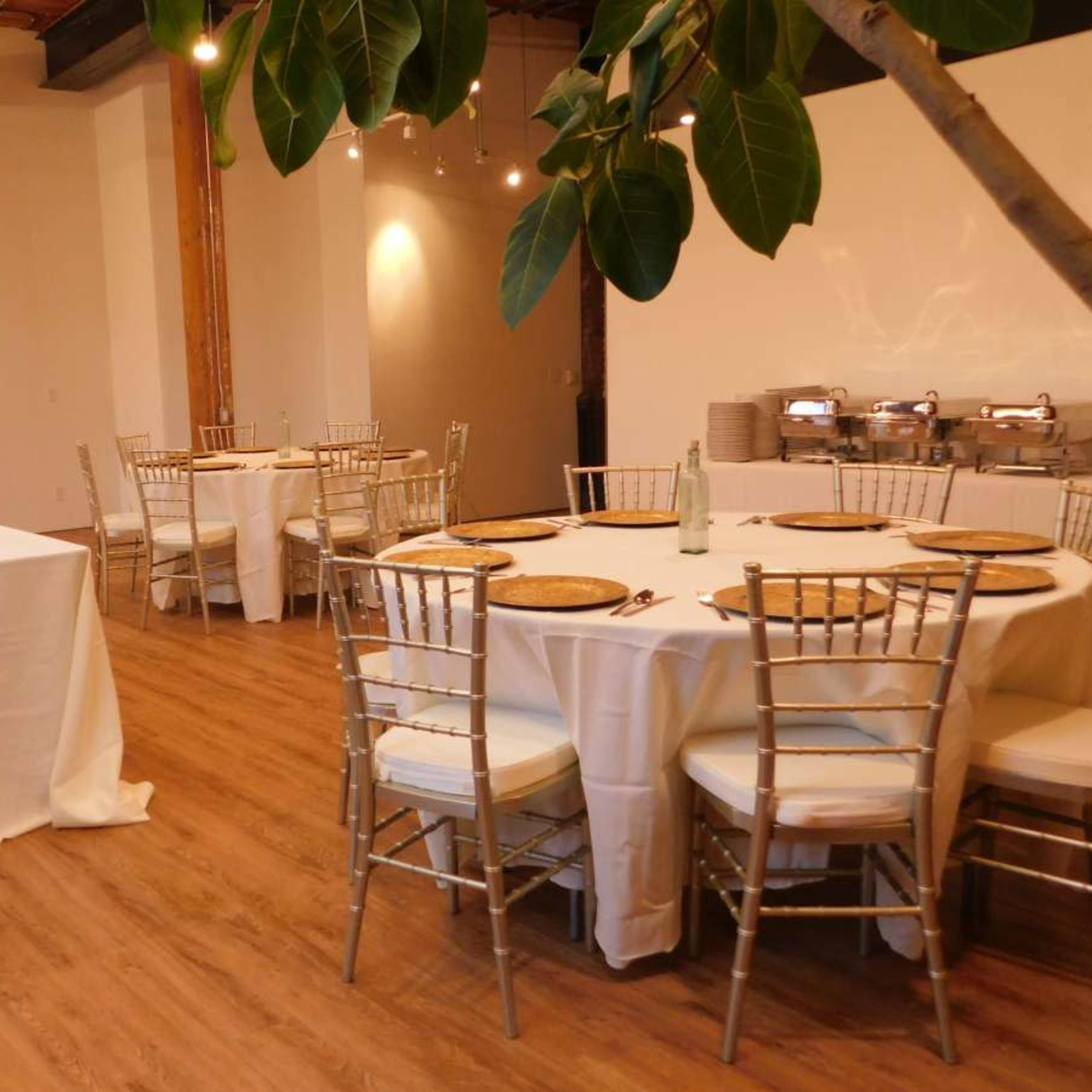 A dining area with several round tables set with white tablecloths and gold charger plates, surrounded by a few potted trees.