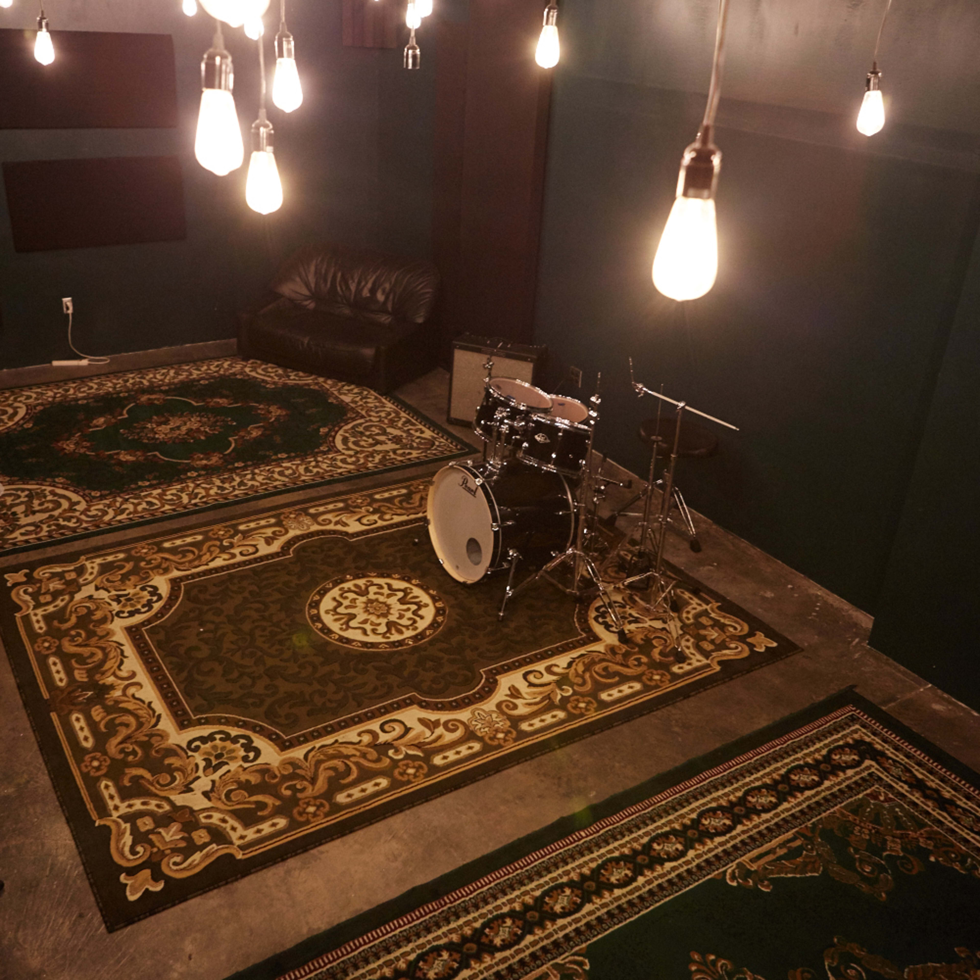 A music studio with a black couch, a drum set, and several hanging light bulbs above patterned rugs on the floor.
