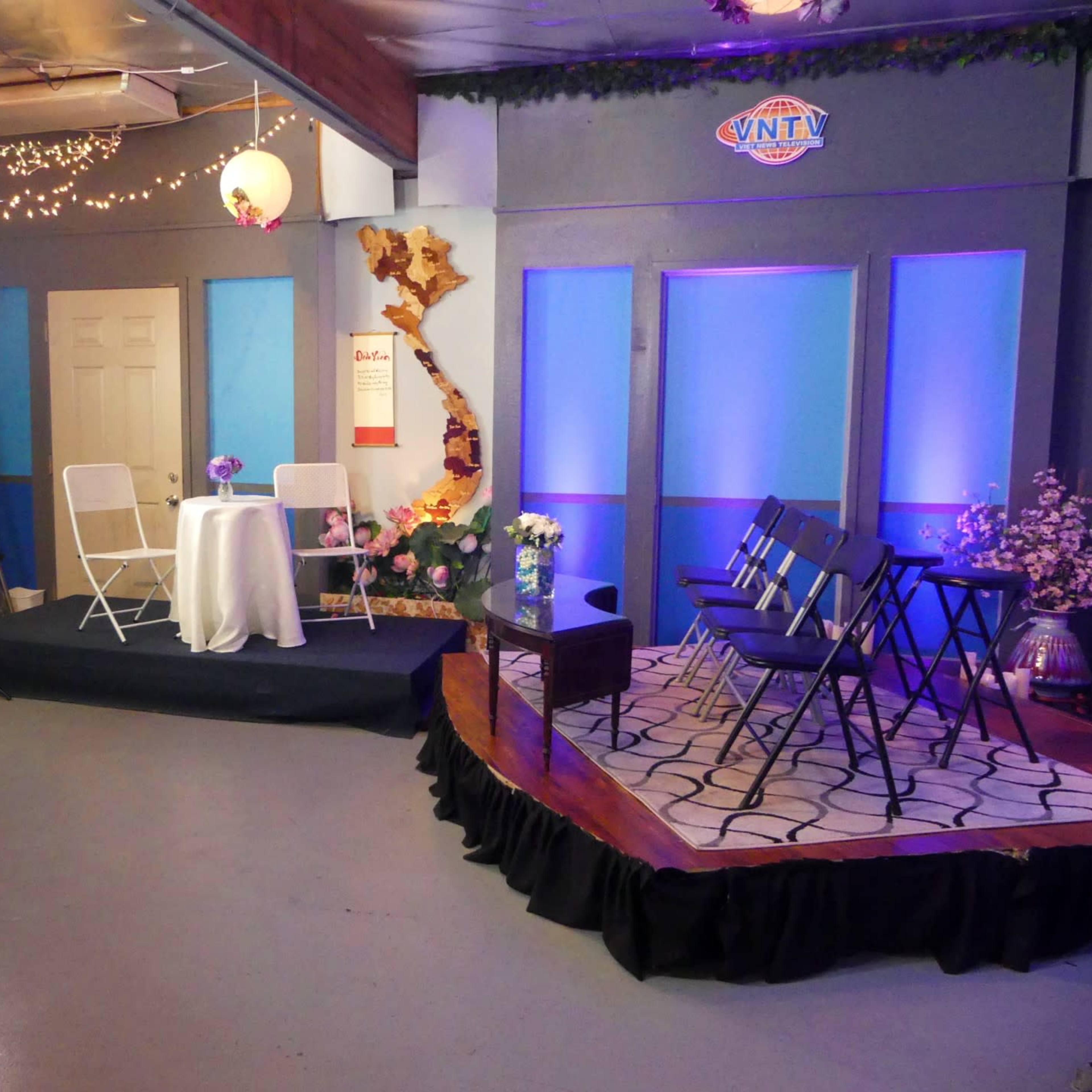 A television studio setup with chairs arranged in a semi-circle around a small table, decorated with flowers and soft lighting.
