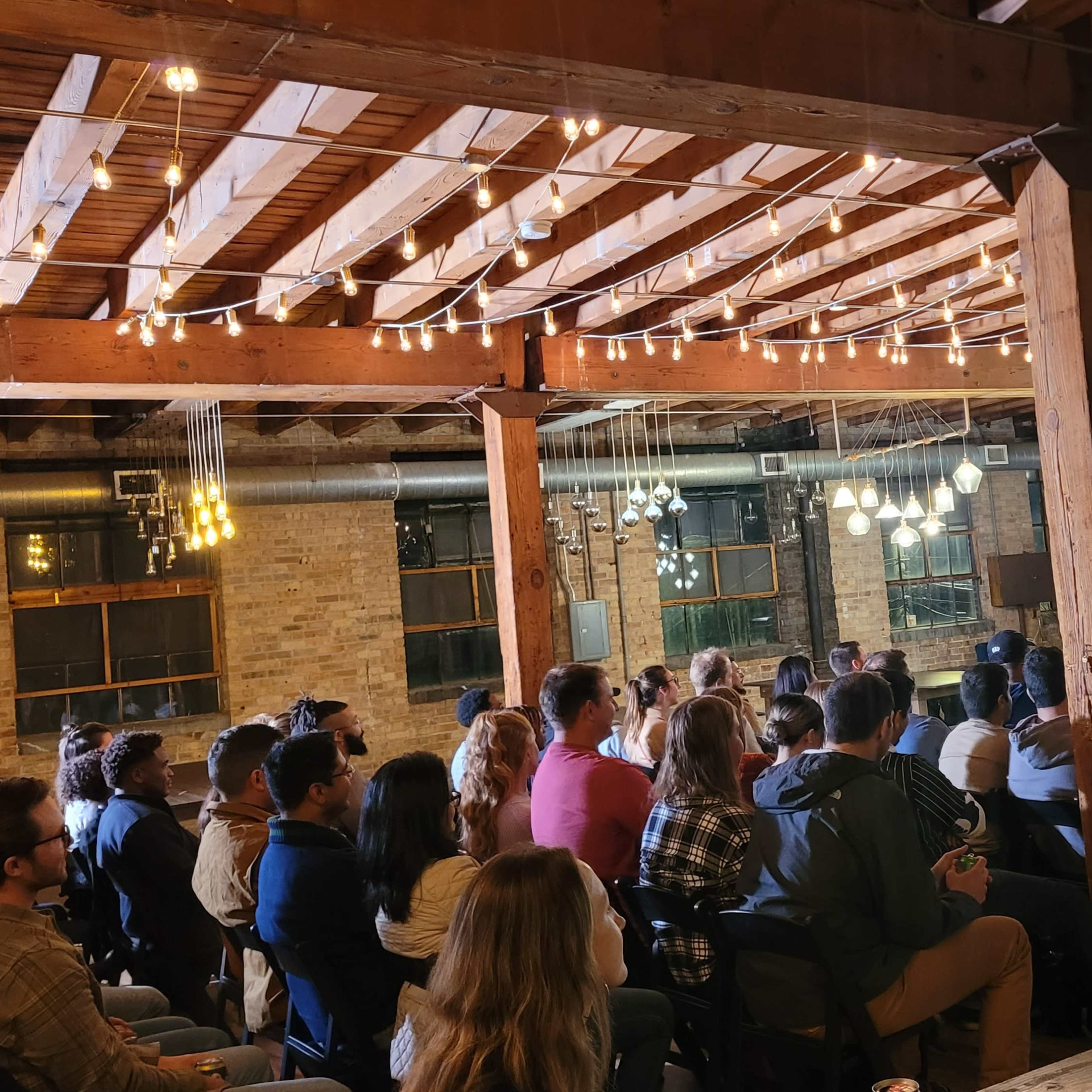 A diverse audience sits in rows facing a speaker on a stage in a rustic venue illuminated by string lights.