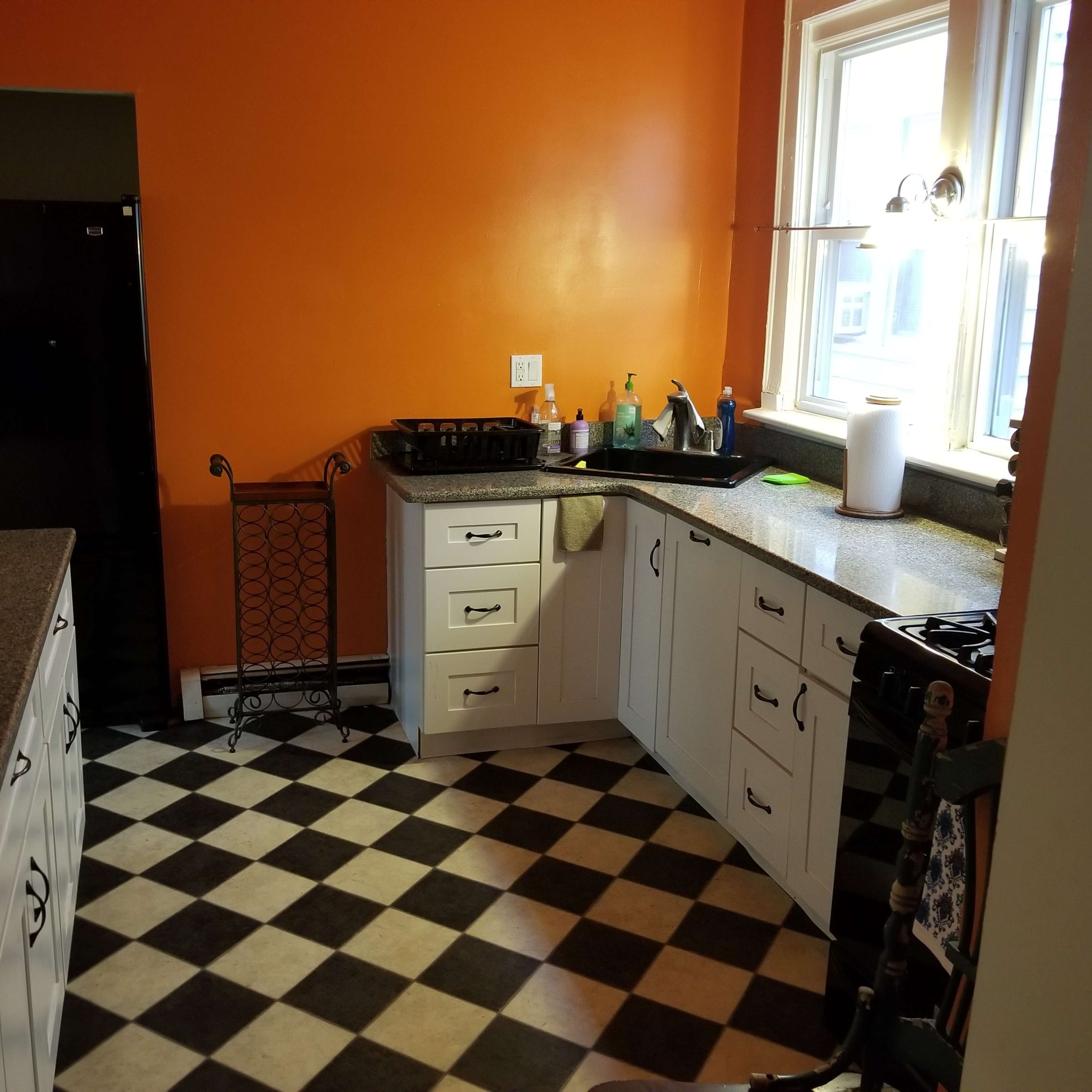A kitchen with orange walls, white cabinets, a black fridge, and a checkered black and white floor.