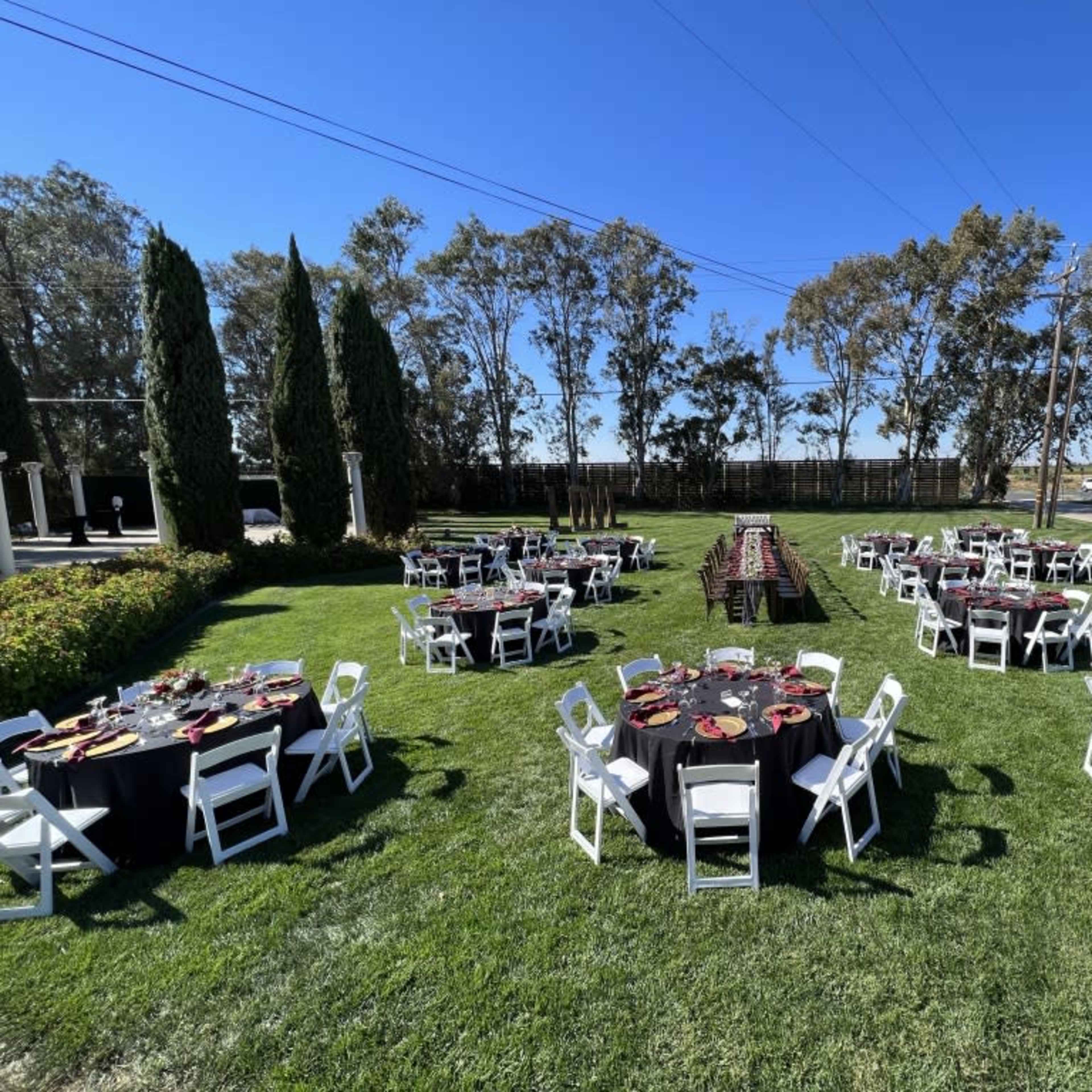 A collection of round tables set for an outdoor event is arranged on a grassy area, surrounded by trees and clear blue skies.