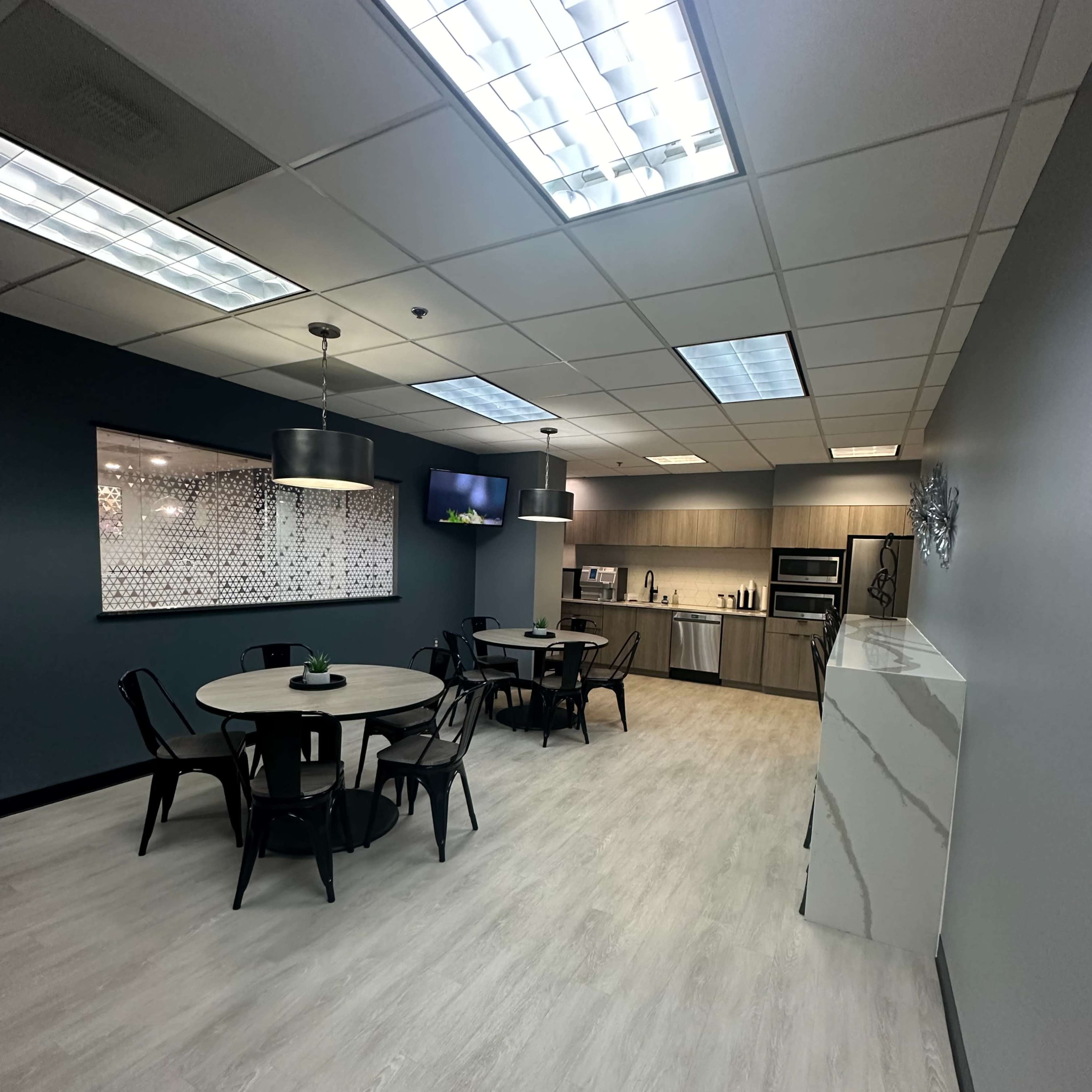 A modern office break room featuring a dining area with a circular table and black chairs, alongside a kitchenette with appliances and a television on the wall.