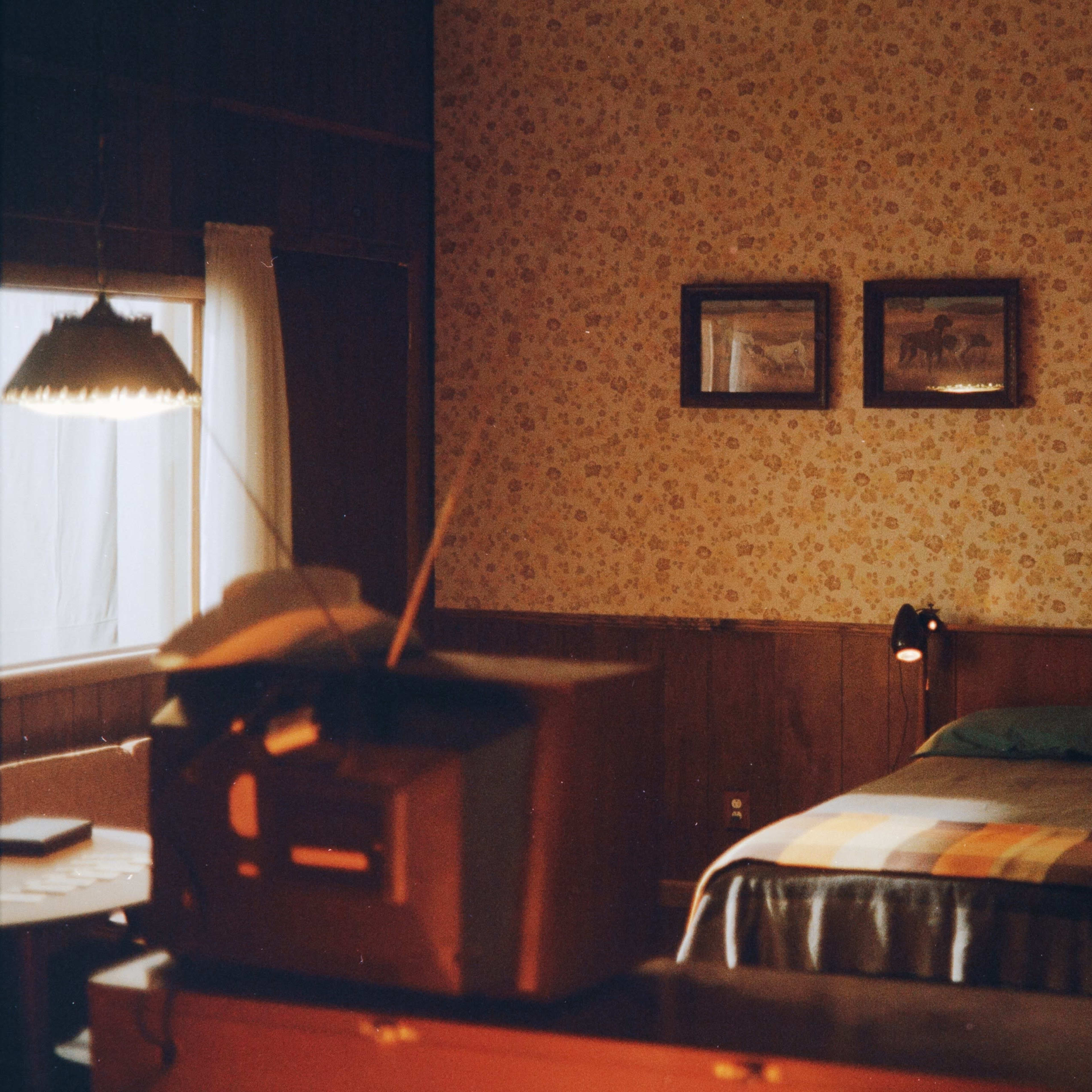 A bedroom with floral wallpaper, a bed covered with a plaid blanket, framed pictures on the wall, and a vintage television set on a dresser.