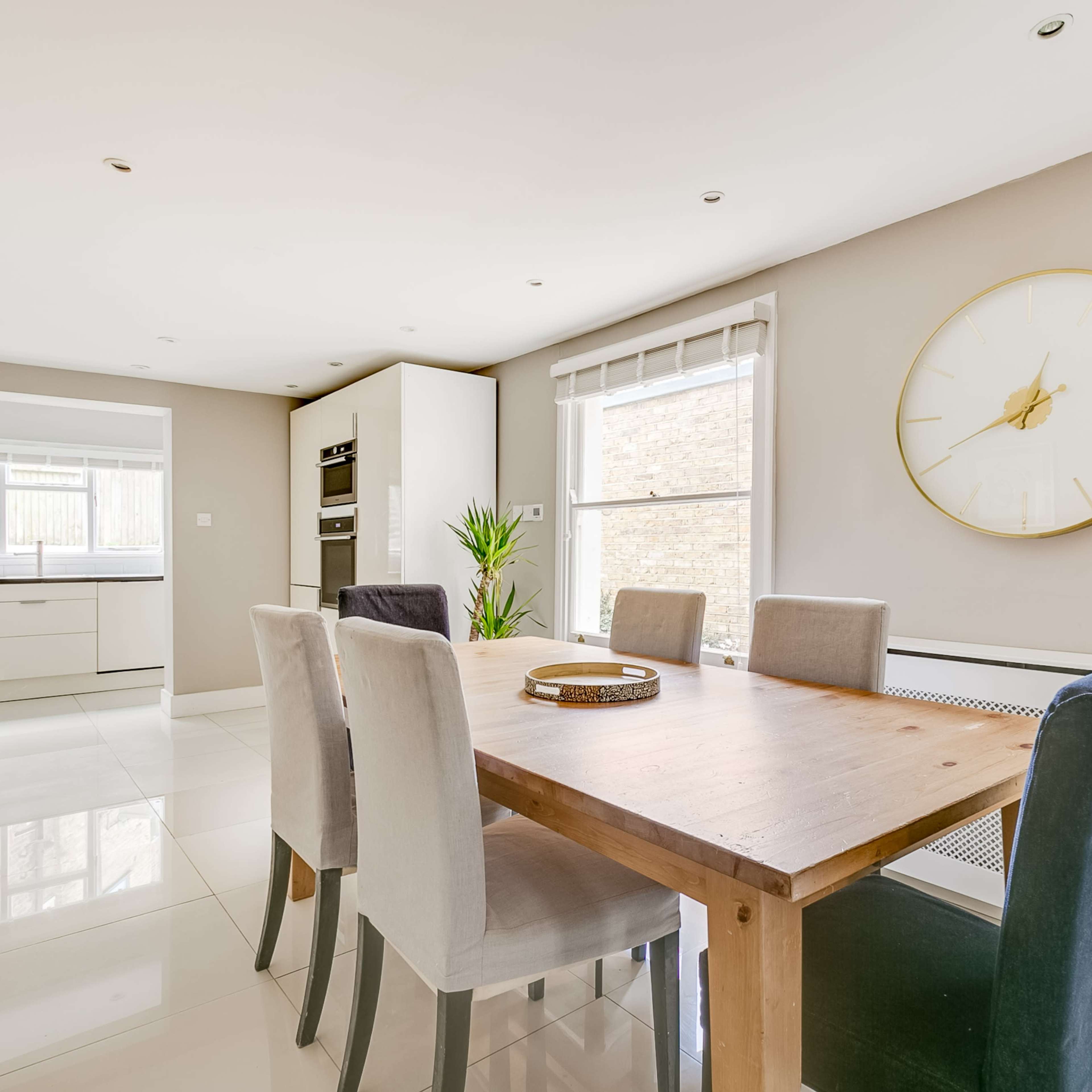 A modern kitchen features a wooden dining table surrounded by chairs, with a large clock on the wall and a sunny window.